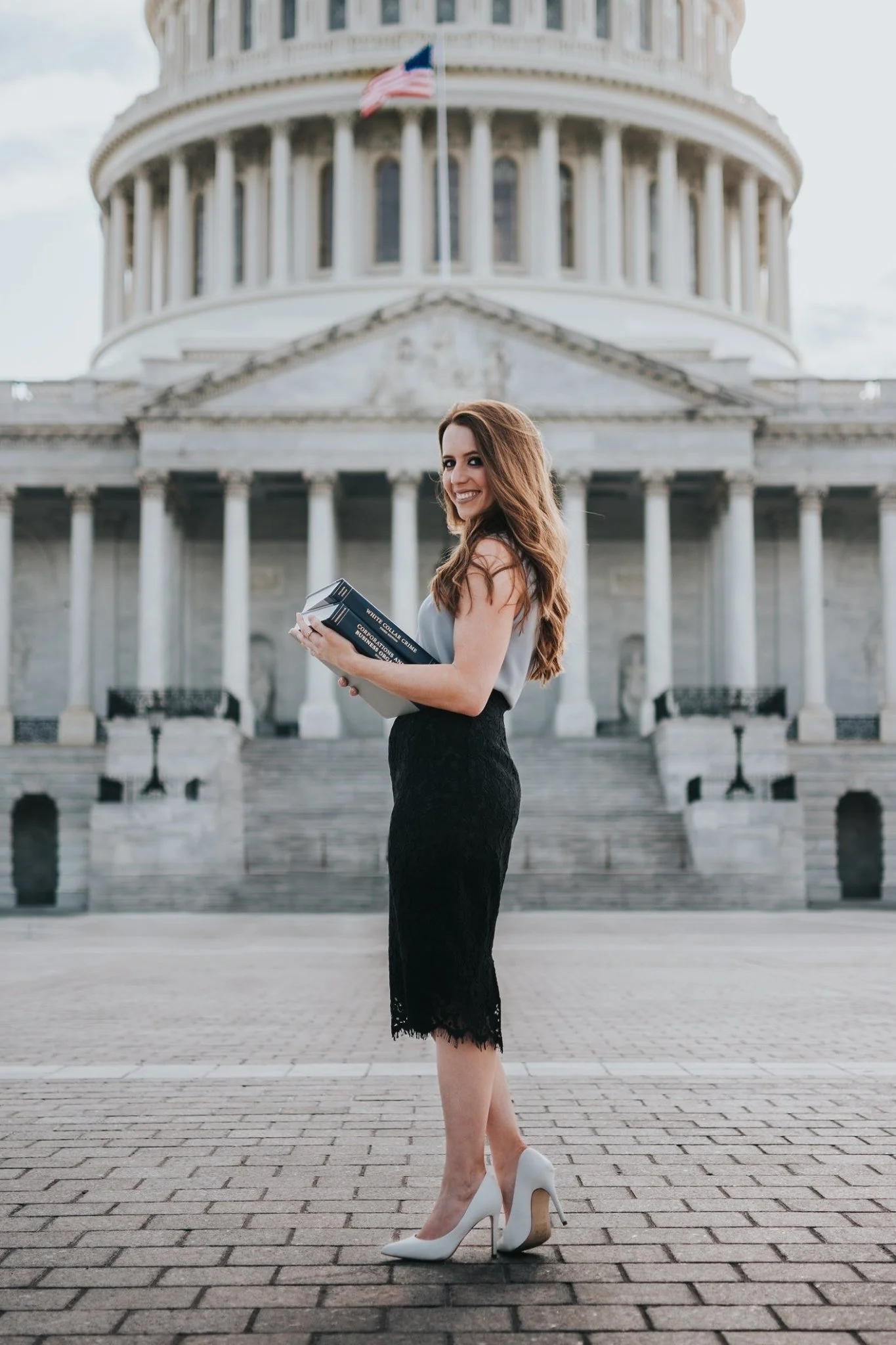 A female artist in a gray blouse, black lace skirt, and white high heels standing outside the U.S. Capitol building, holding a law text book and smiling at the camera.