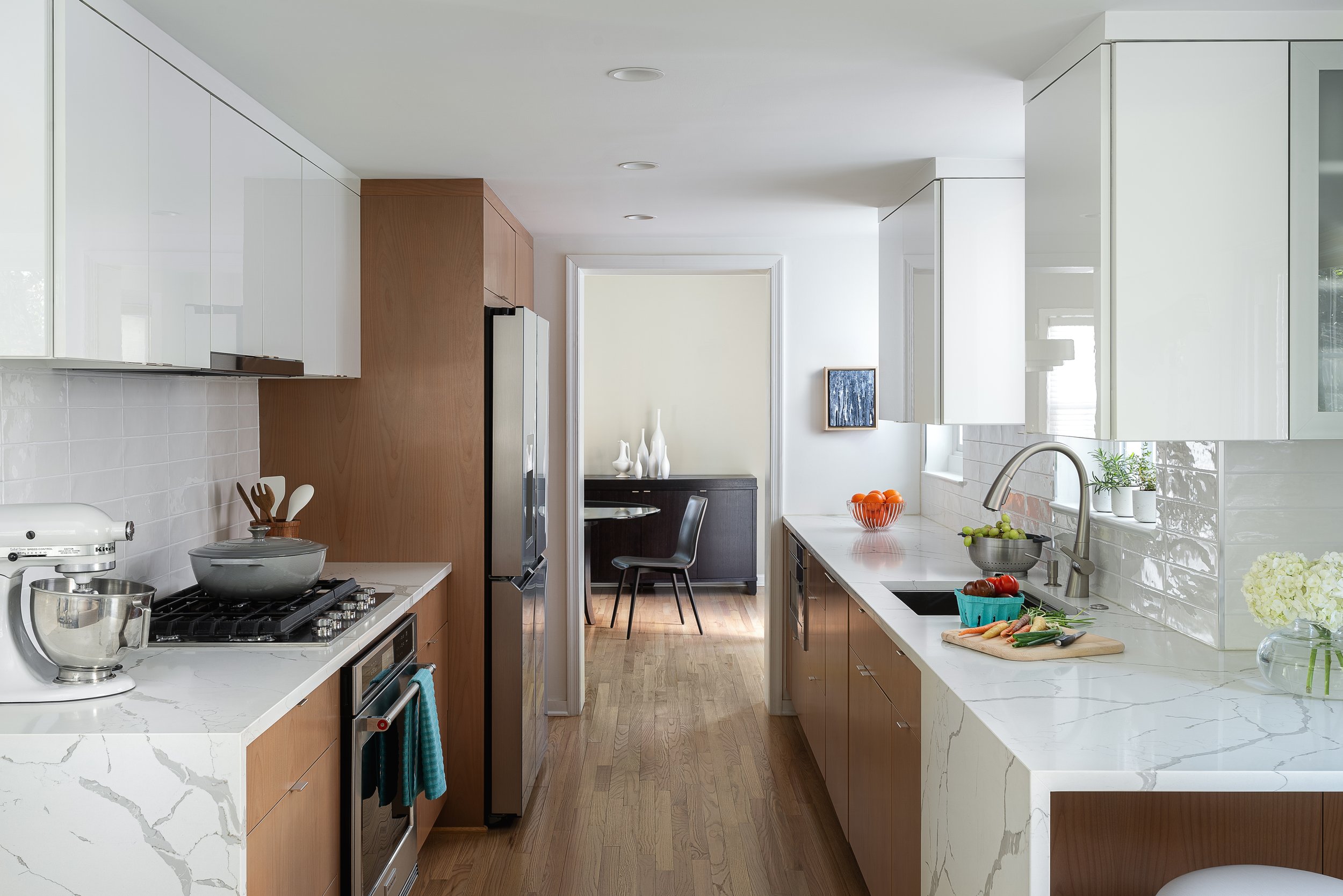Modern kitchen with white marble countertops, wooden lower cabinets, white upper cabinets, and stainless steel appliances, featuring an original framed painting on canvas in the background.