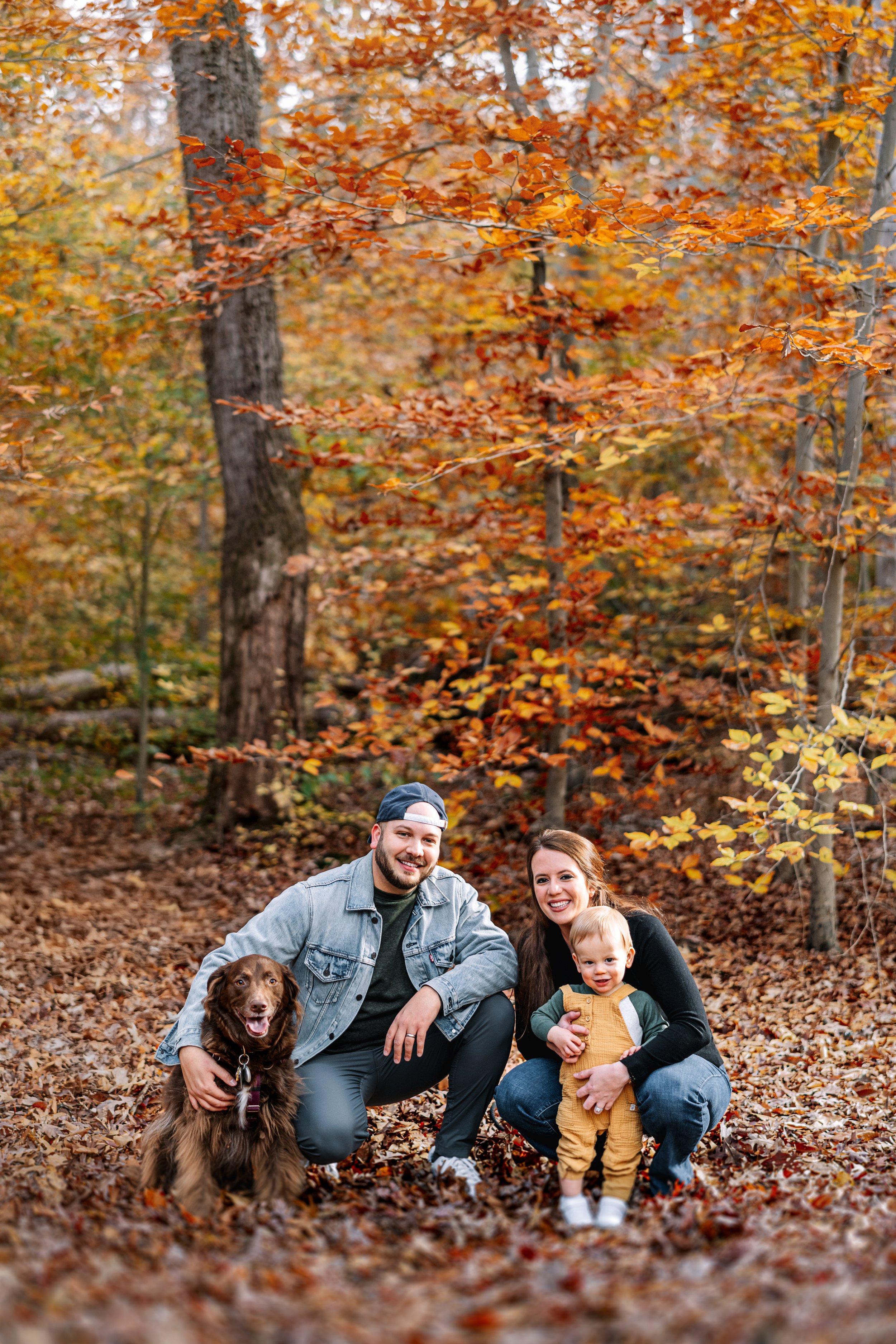 A happy family of three with a dog in a fall forest with orange and yellow leaves. The family includes a man, a woman, and a young child in mustard overalls. The man is wearing a denim jacket and a cap; the woman has long brown hair; the child has blonde hair and is smiling. The dog is a brown retriever sitting beside the man.
