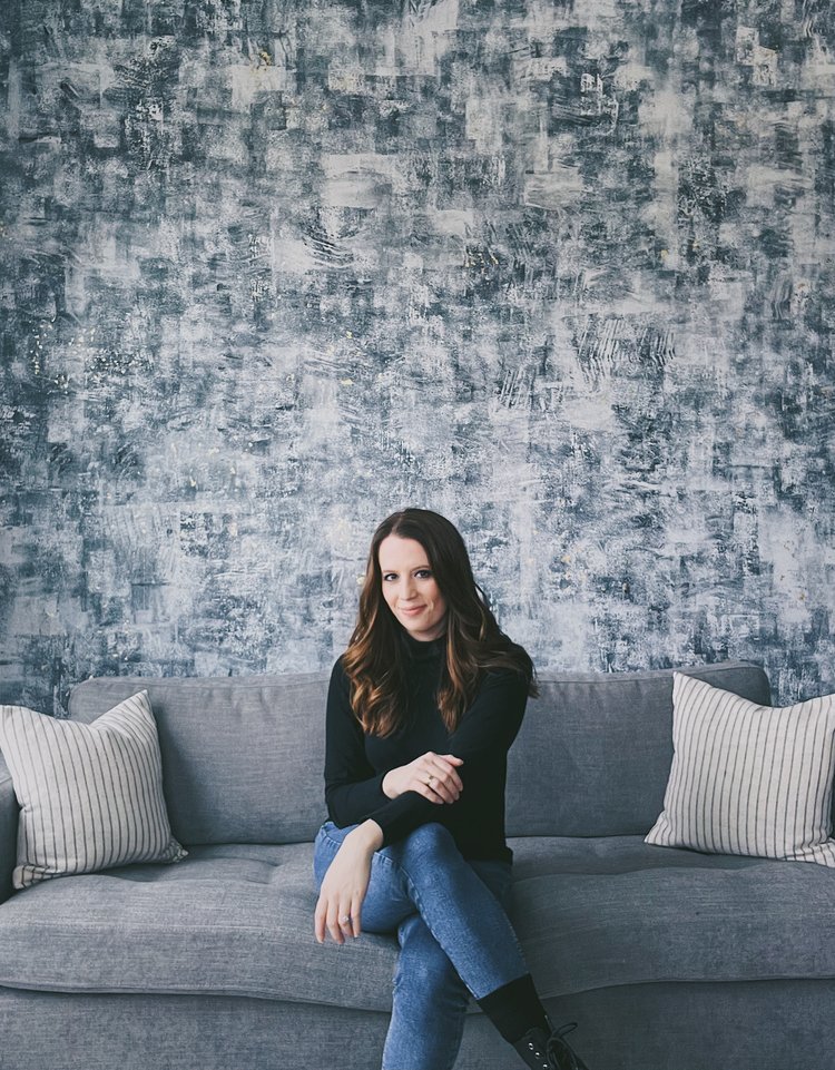 Female DC artist with long brown hair in a black top and jeans sitting on a gray sofa against an original abstract painted blue-gray mural wall, smiling and crossing her legs.