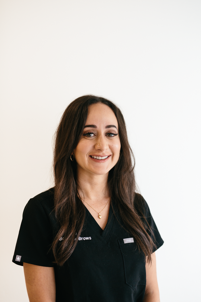 Young woman with brown hair wearing pink medical scrubs standing against plain light-colored background.