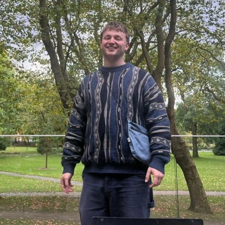 A smiling man wearing a patterned sweater and dark pants standing outdoors in a park with trees in the background.