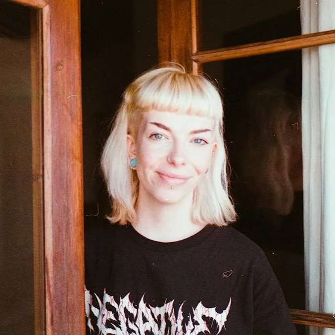 Young woman with blonde hair and blue earrings smiling near a wooden window frame.