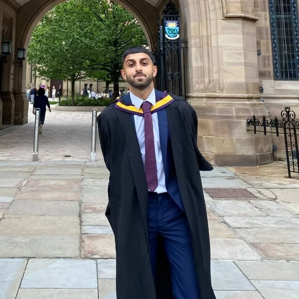 A man dressed in a graduation gown and cap standing outdoors in front of a university building with arches and a shield emblem.