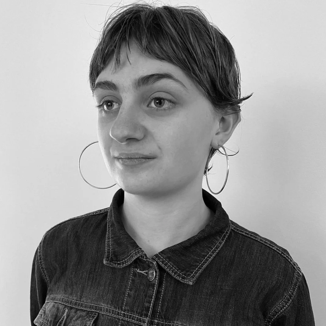 Black and white portrait of a young woman with short hair, wearing hoop earrings and a denim jacket, looking slightly to the side.