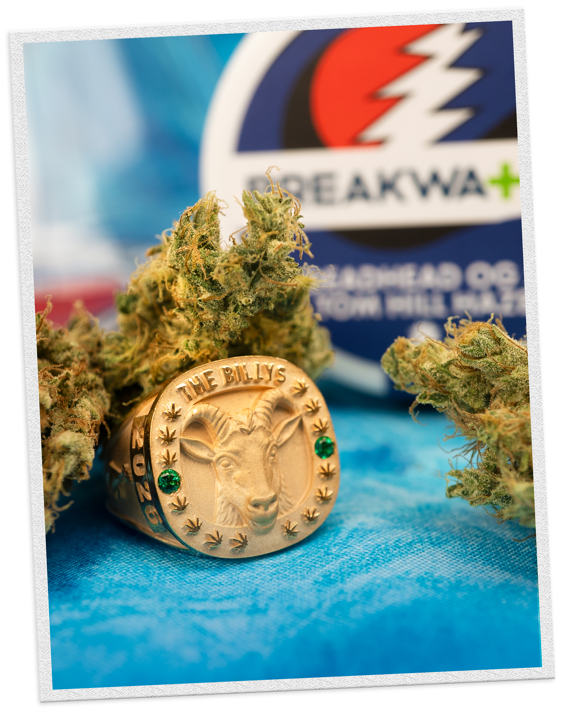 Gold Billy's ring with green stones and a goat head with cannabis and Deadhead OG x Tom Hill Haze packaging in the background