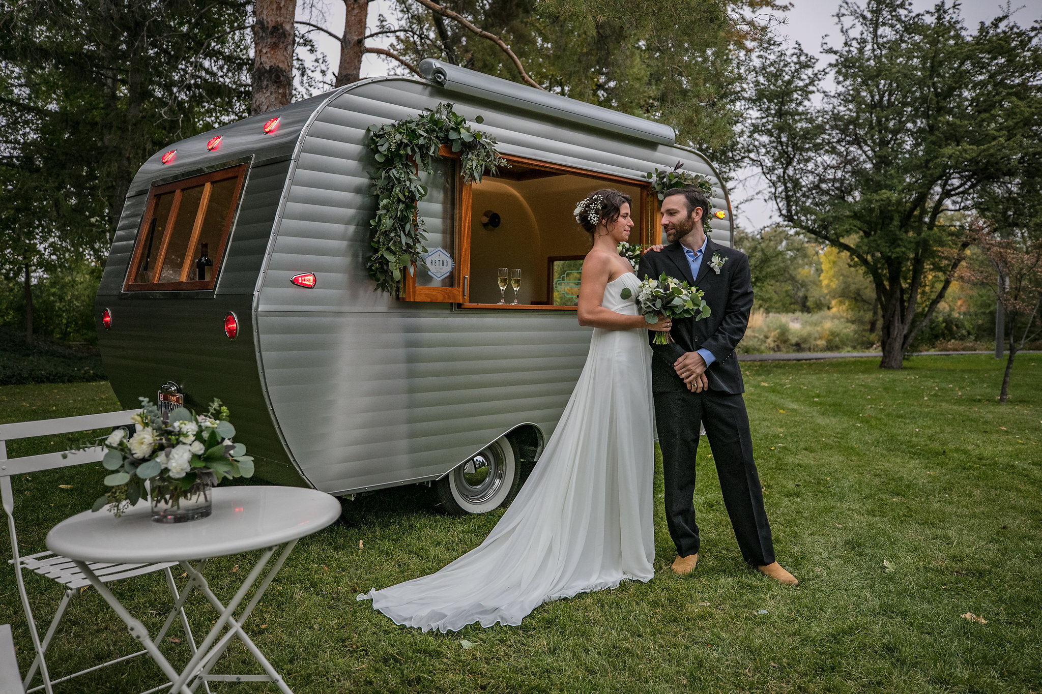 Bride and groom standing outside a silver vintage camper decorated with greenery, with a small white table and flower arrangement nearby, on a grassy area with trees in the background.