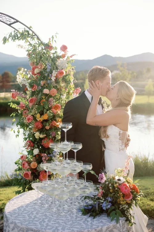 A newlywed couple shares a kiss at their outdoor wedding reception near a lake, with a floral arch and a champagne tower in the foreground.