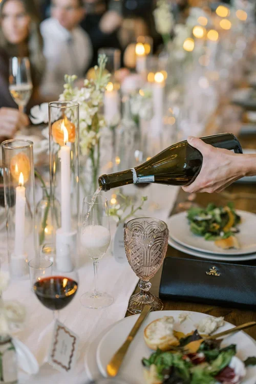 Person pouring champagne into a glass at an elegantly set dinner table with candles and floral arrangements.