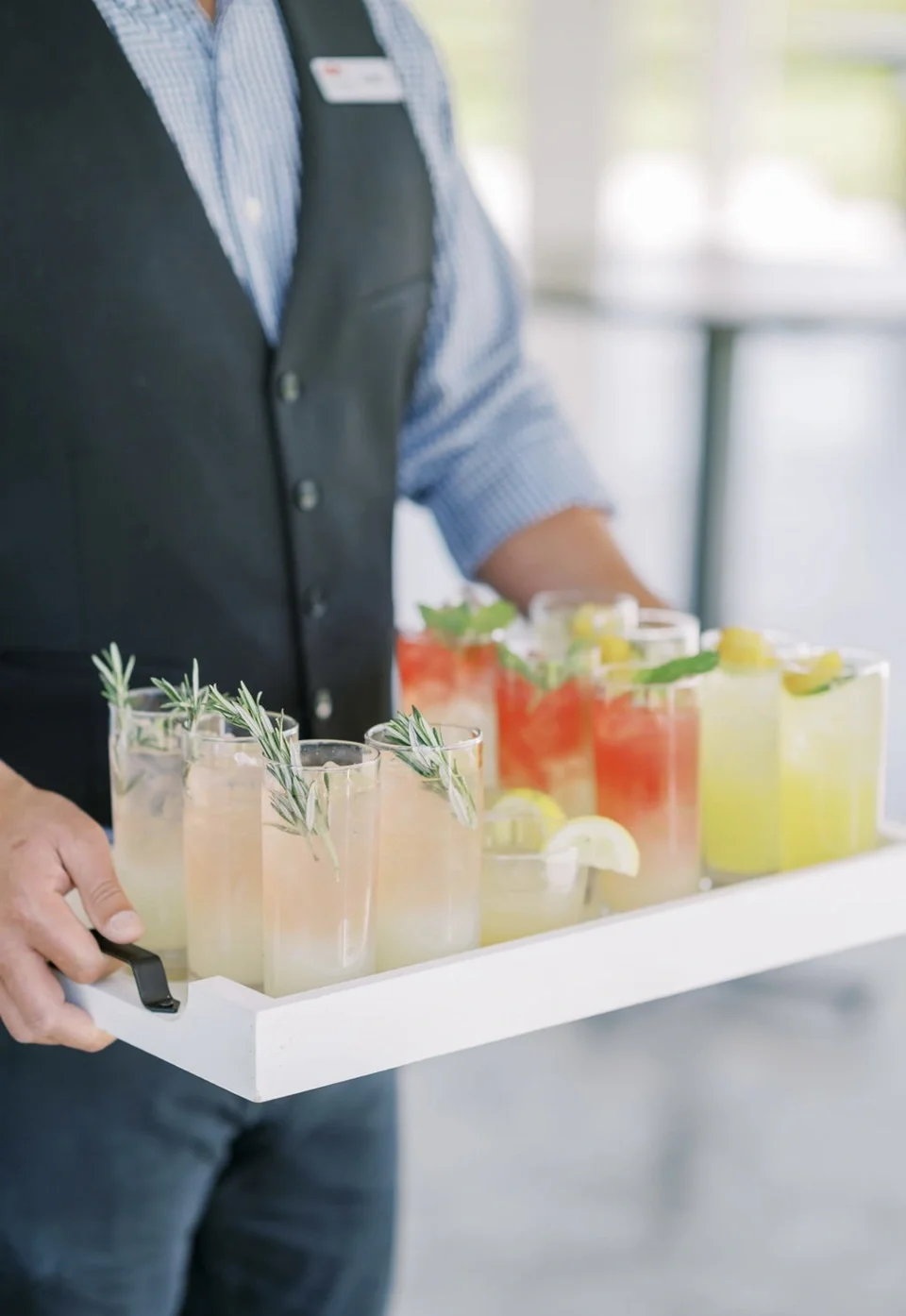 A person holding a tray of colorful beverages garnished with herbs and lemon slices, at a social event or gathering.