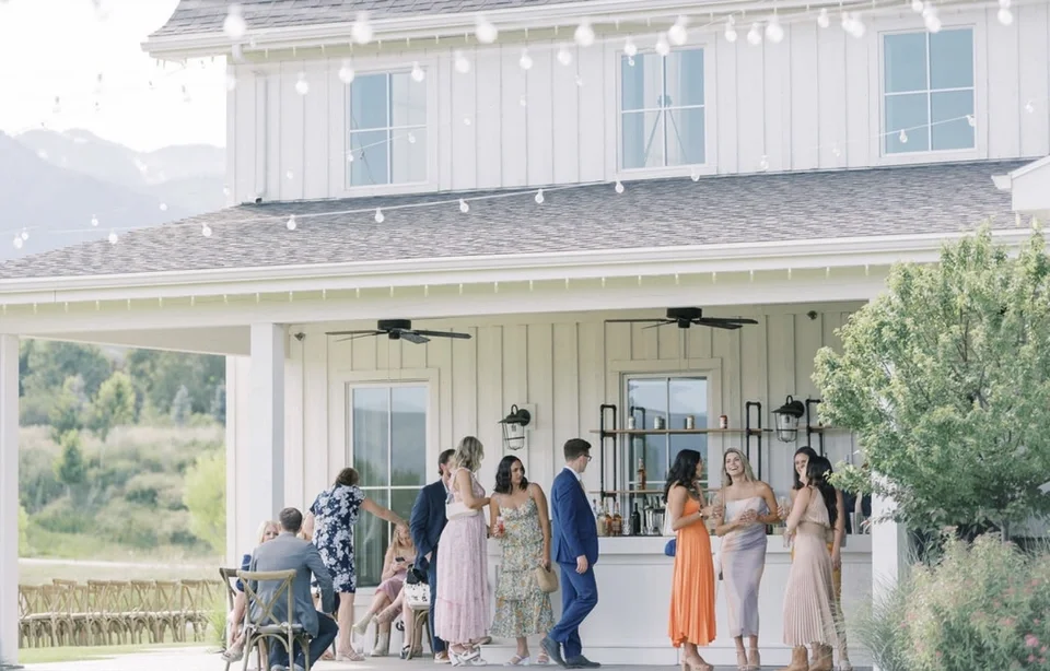 People gathering and socializing at an outdoor party or event on a patio with string lights, in front of a white house with a bar area.