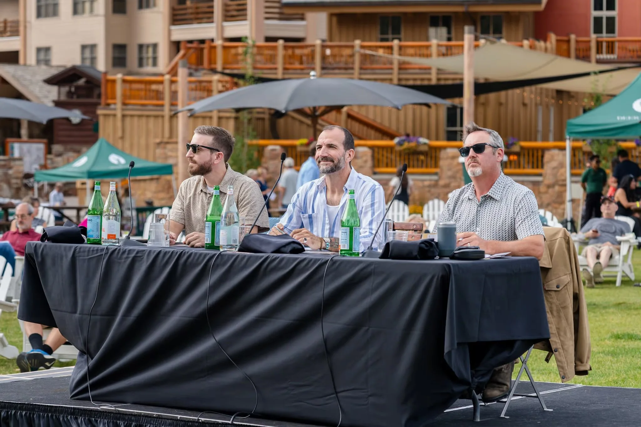 Three men sitting at a table outdoors, possibly at a panel or discussion event, with bottles of water in front of them and a lush background featuring buildings, umbrellas, and people.