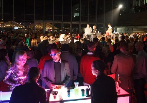 A crowd gathered at an indoor concert or event with a stage featuring performers, and audience members seated or standing, some illuminated by colorful lights.