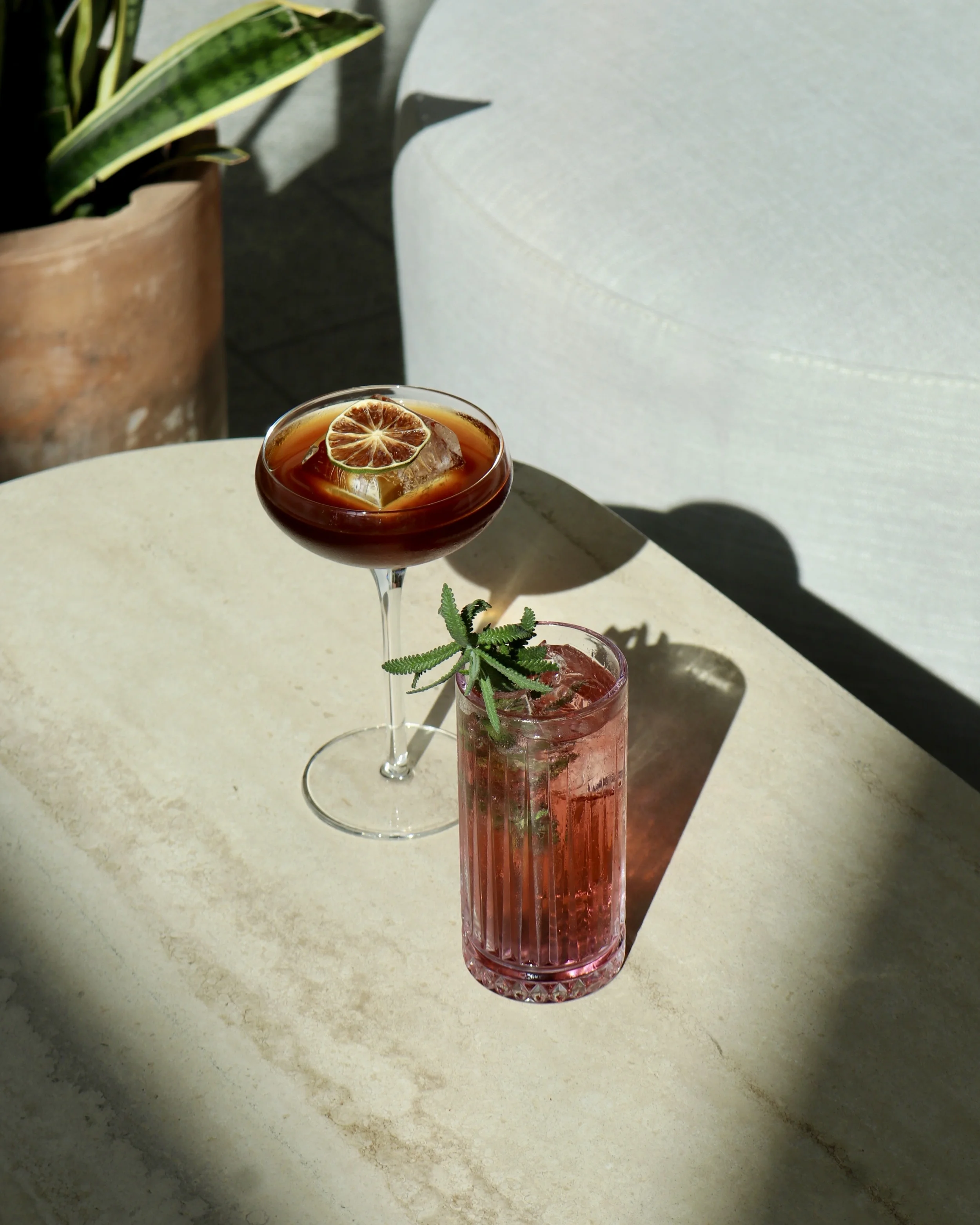 Two cocktails on a round beige table; a dark brown drink with a dried citrus slice and ice in a coupe glass, and a pink drink garnished with a sprig of greenery in a tall ribbed glass; sunlight casting shadows.