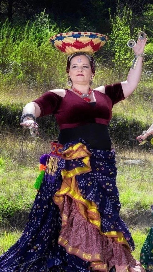 A woman dressed in colorful, layered clothing with an elaborate skirt, wearing jewelry, standing outdoors with trees and grass in the background. She balances a decorated basket on her head and holds a pair of googly eyes in one hand.