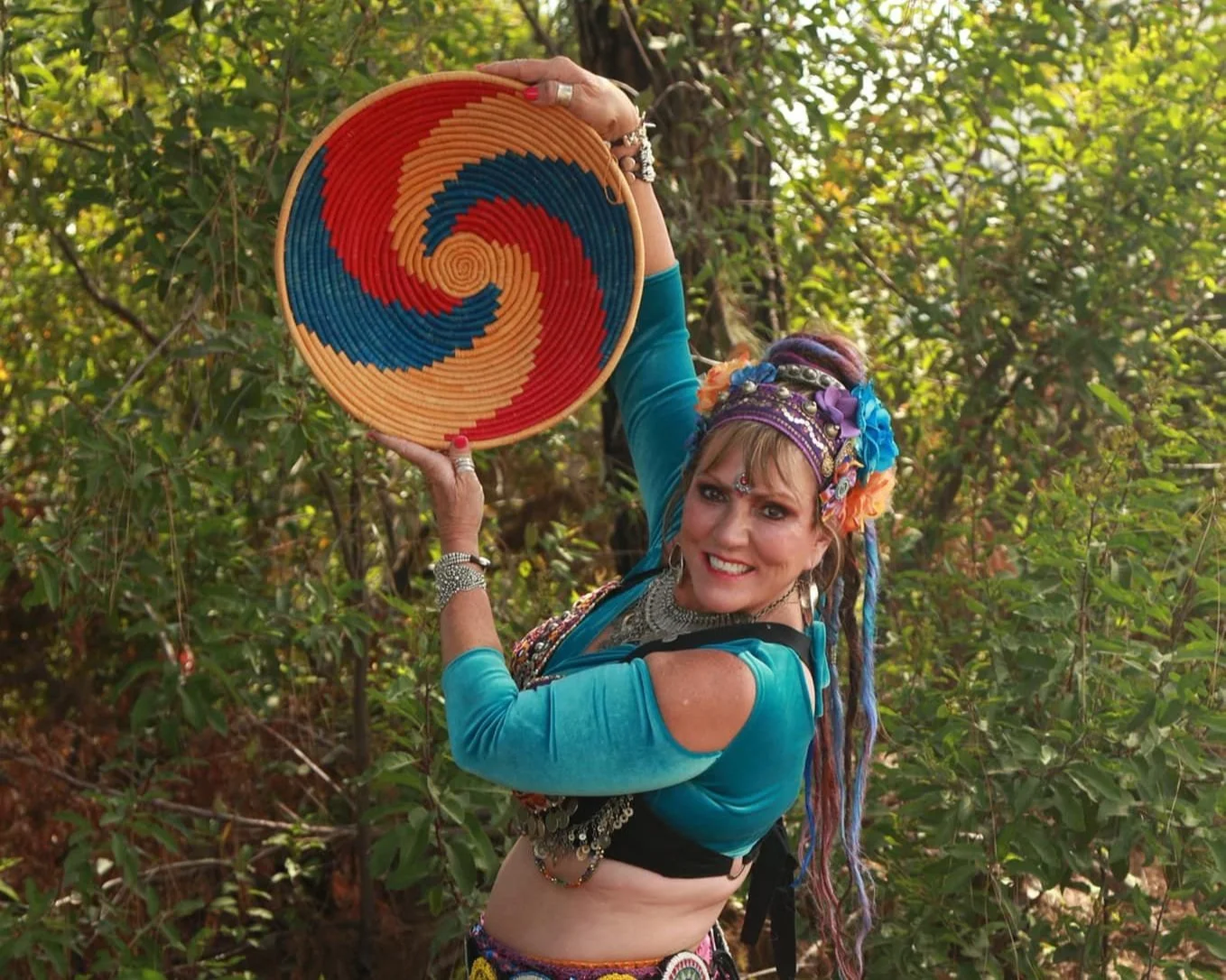 Woman in colorful clothing and accessories posing outdoors with a woven circular artwork featuring a spiral pattern of red, orange, blue, and beige.