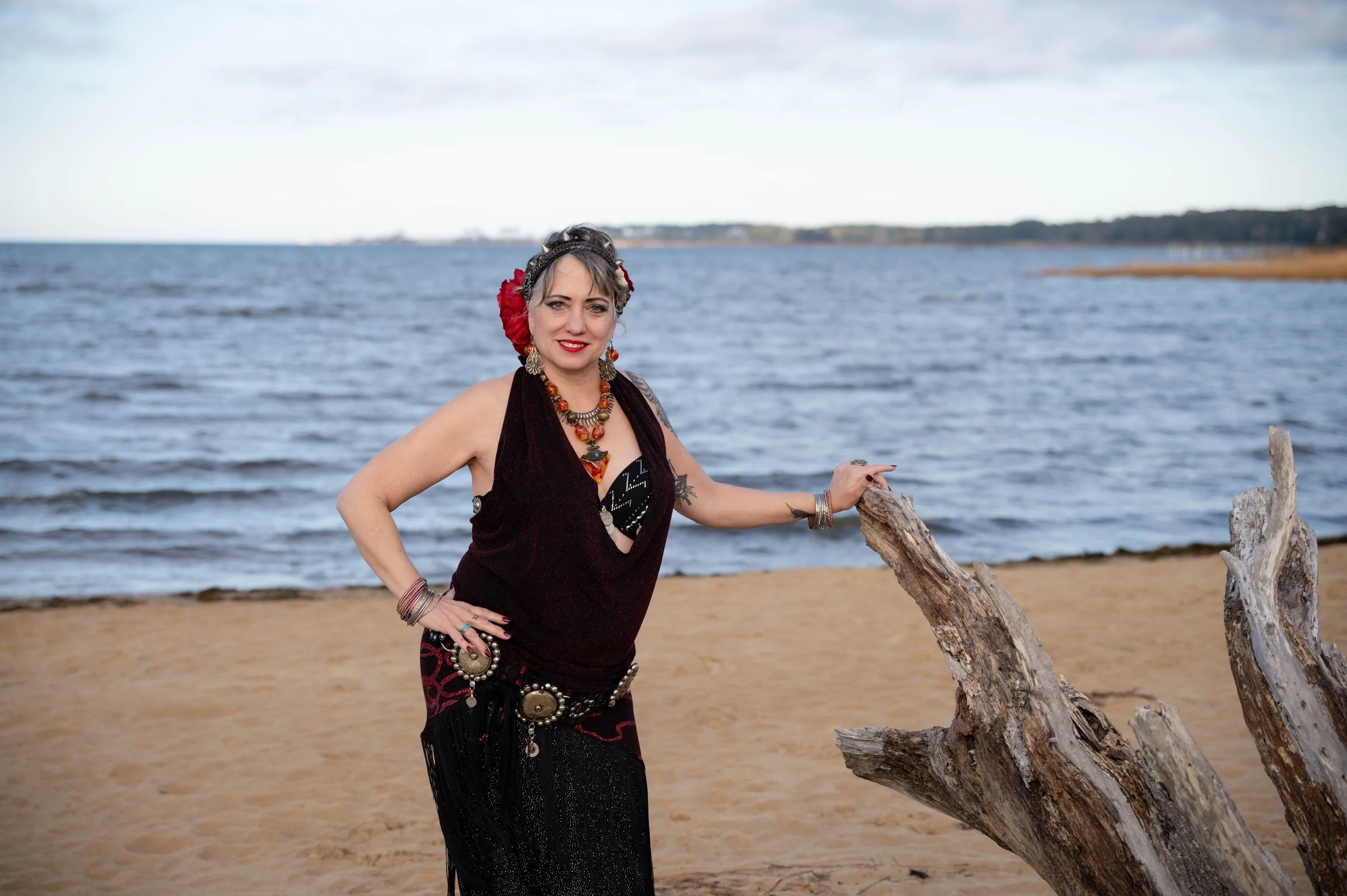 A woman with gray hair, wearing a black dress with jewelry, standing on a sandy beach near a driftwood log, with the ocean and distant shoreline in the background.
