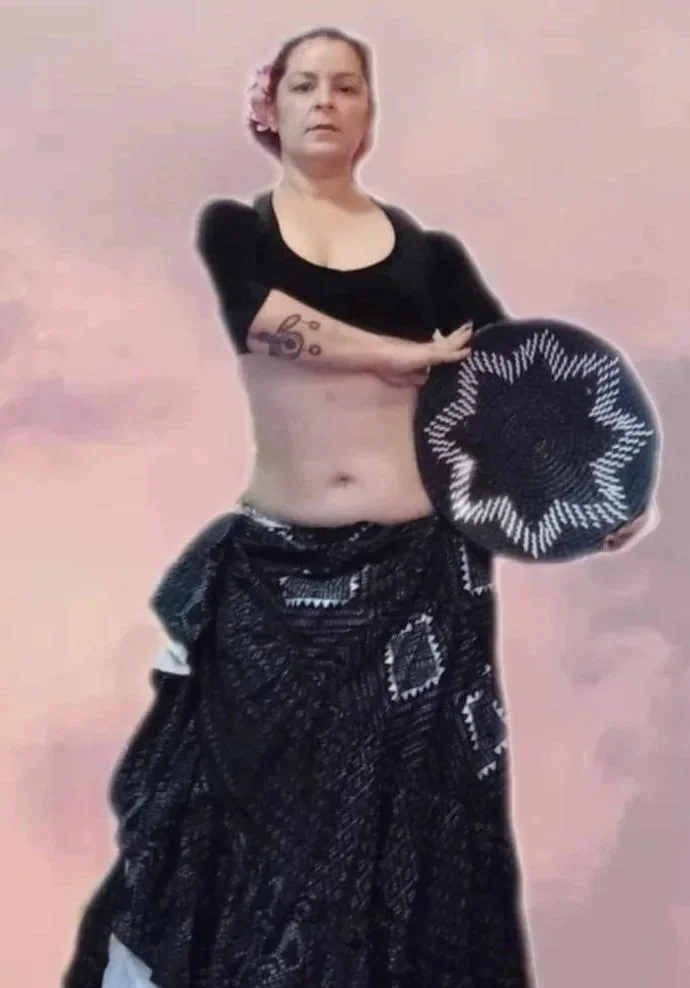 A woman with pink hair, wearing a black top, holding a round woven object, with a black patterned skirt, standing against a plain pink background.
