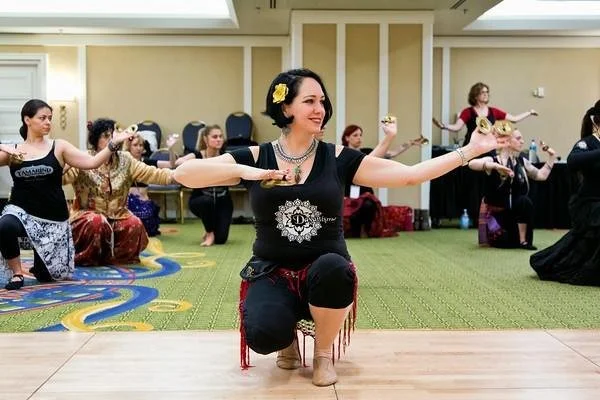 Group of women practicing a traditional dance in a spacious room. One woman in the foreground is smiling, holding a prop, and kneeling, while others behind her are in various dance poses.
