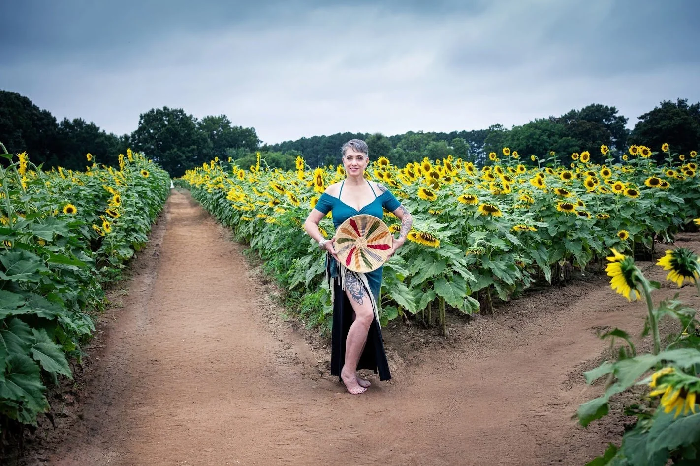 A woman with short gray hair, tattooed arm, wearing a teal off-shoulder dress, standing barefoot in a sunflower field, holding a large round hat, with trees and a cloudy sky in the background.