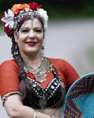 A woman dressed in belly dance attire with a floral headpiece and jewelry, carrying a colorful woven basket.