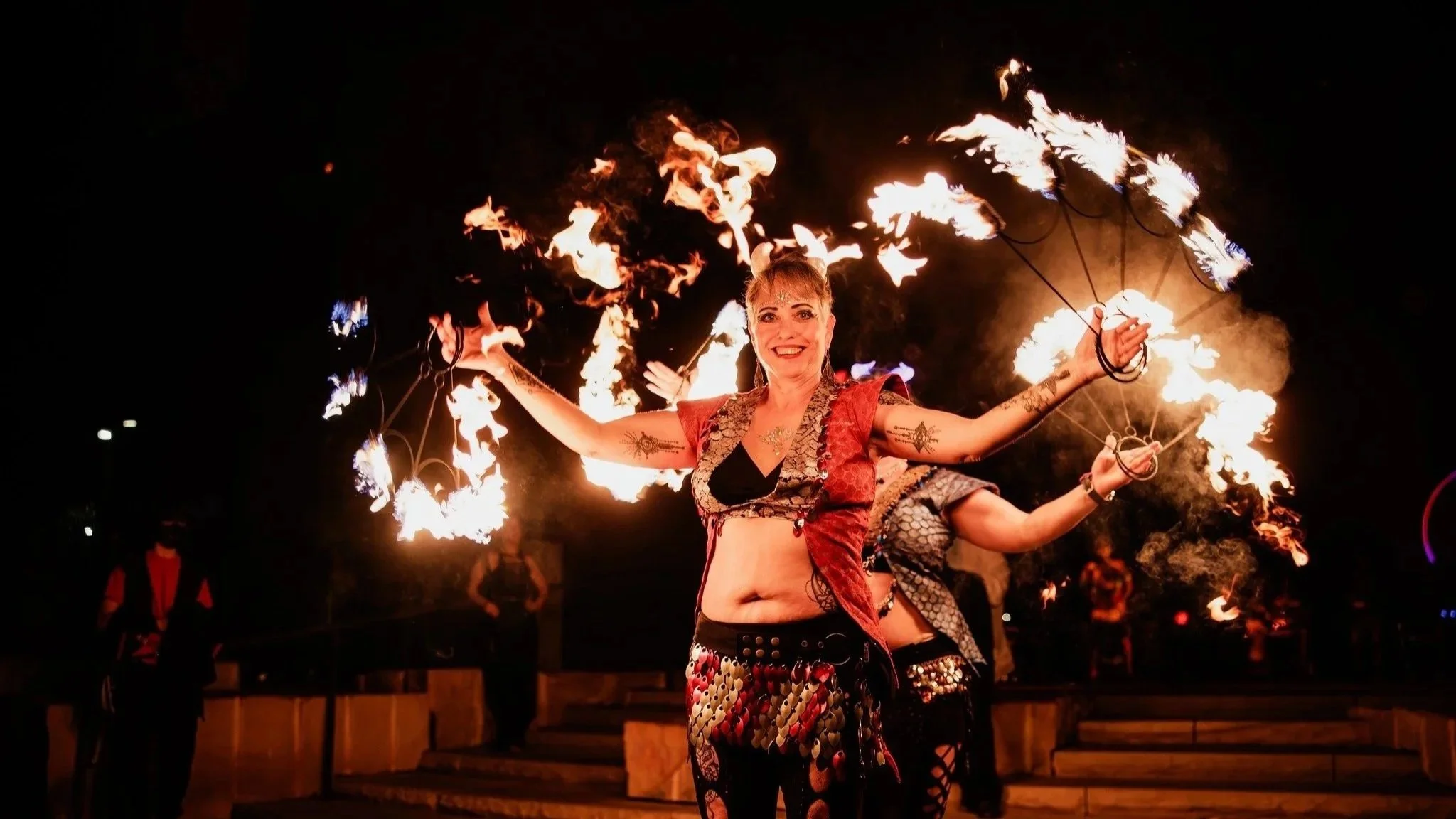 A woman performing a fire dance at night, holding fire poi, with flames in the air and a big smile on her face.
