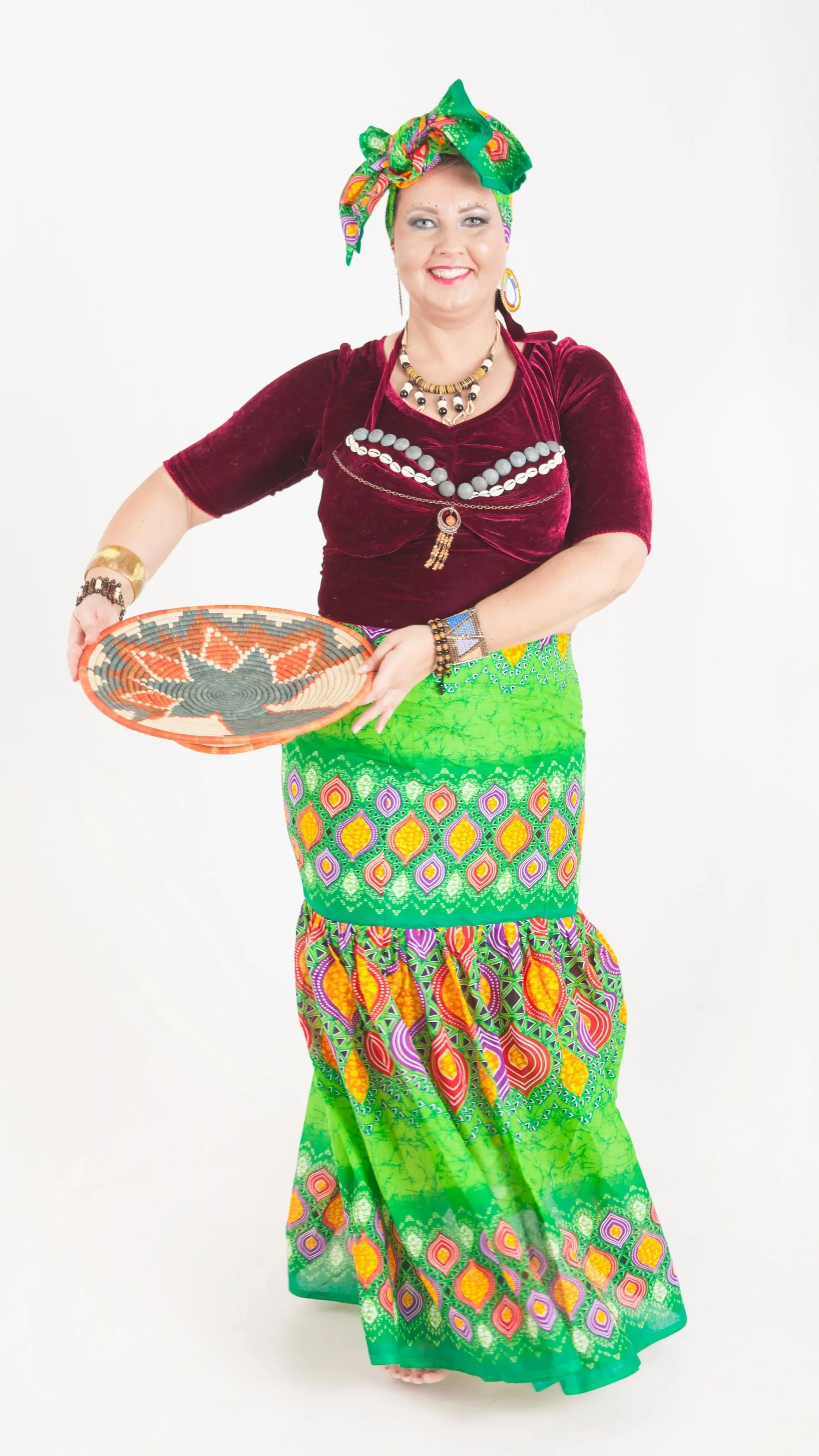 A woman dressed in a colorful costume, holding a woven tray, with bright makeup, jewelry, and a vibrant head wrap, standing against a plain white background.
