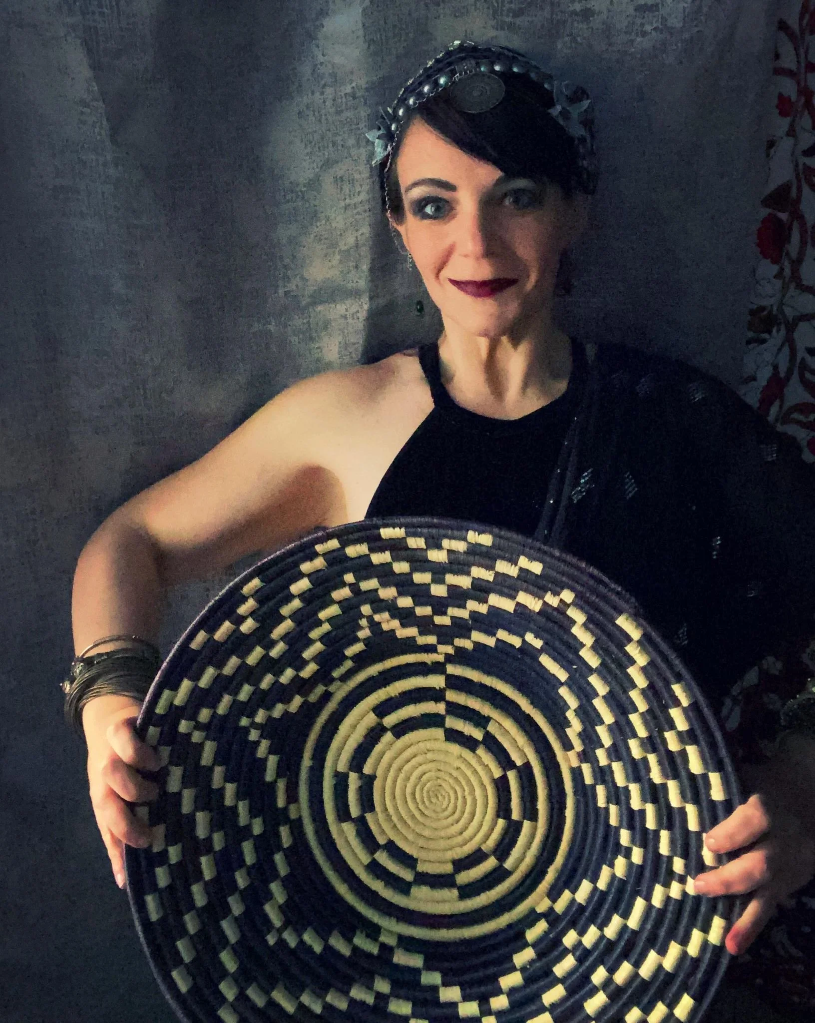 A woman with dark hair and makeup holding a large, round, woven basket with a black and yellow spiral pattern, standing against a dark wall.