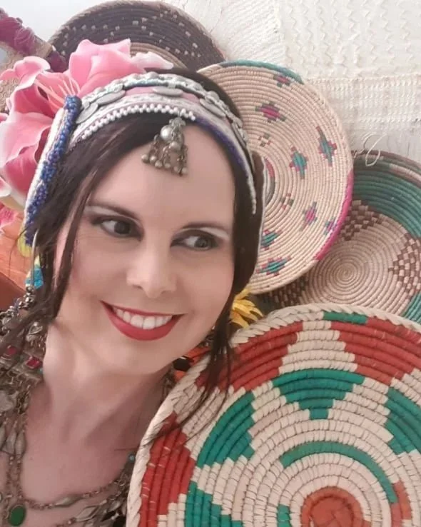 A woman smiling at the camera surrounded by colorful woven baskets and flowers, wearing traditional headwear and jewelry.