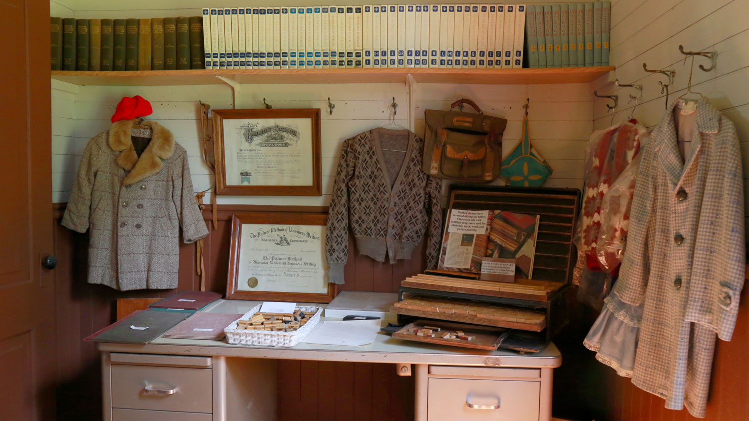 A display with a teacher's desk, clothing and school supplies of bygone eras