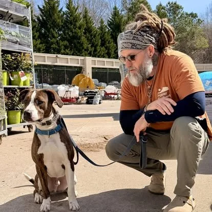 A man with a beard, dreadlocks, and sunglasses crouches next to a brown and white puppy outdoors in a yard, both on a leash.