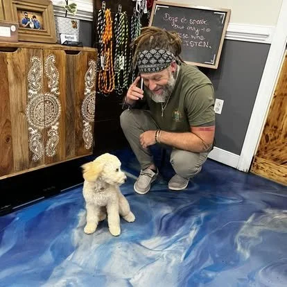 A man with long hair, a beard, and a patterned headband squatting next to a small white dog with fluffy fur. They are indoors, with wooden and black furniture, colorful leashes or collars hanging on the wall, and a chalkboard in the background.
