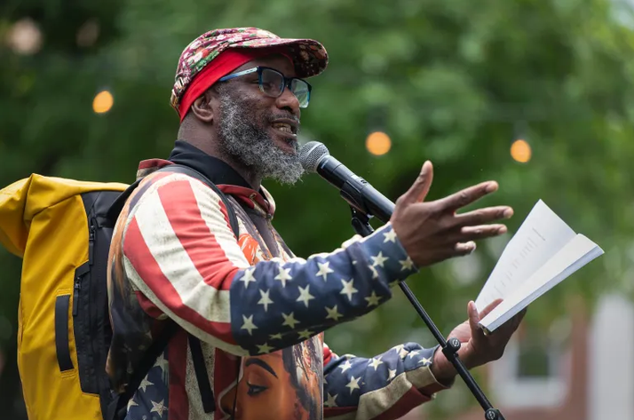 Rajnii Eddins, reciting poetry while standing in front of a microphone and holding a book.