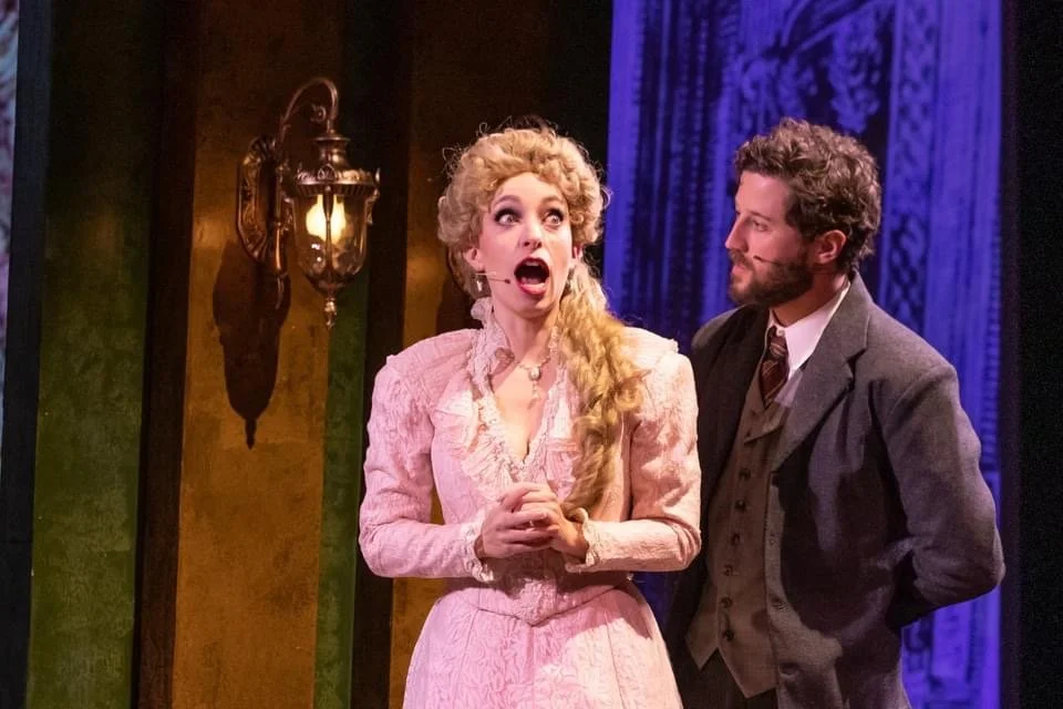  Leslie as Sibella Hallward, wearing pink dress and blond wig, in  A Gentleman’s Guide to Love and Murder  with Lamplighters Music Theatre, pictured with castmate. Photo by Joe Giammarco. 