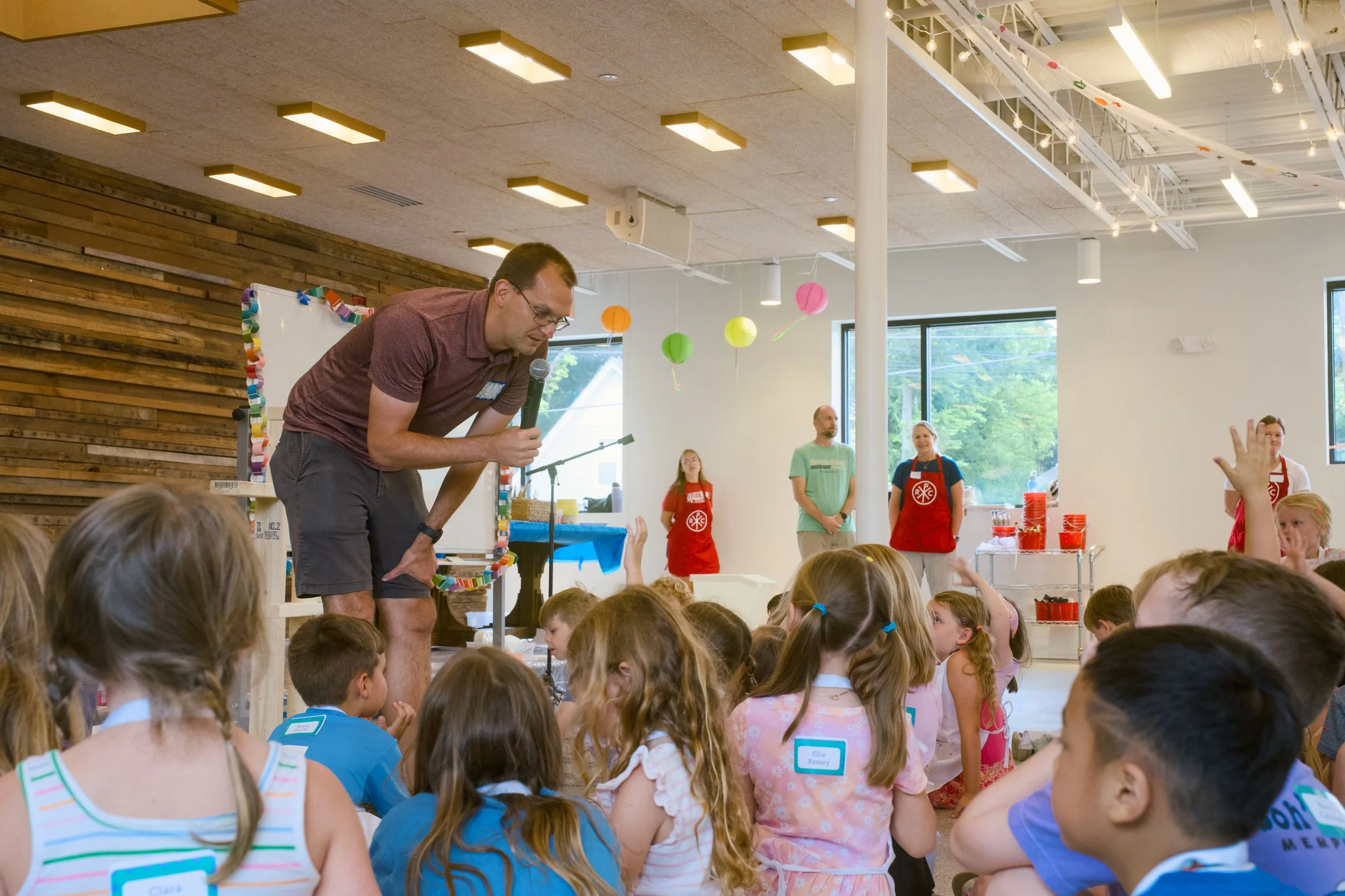 A man with glasses bending down holds a microphone in his right hand while engaging with children seated on the floor during an indoor event. Several adults are in the background, with some wearing red aprons, and colorful decorations hang from the c