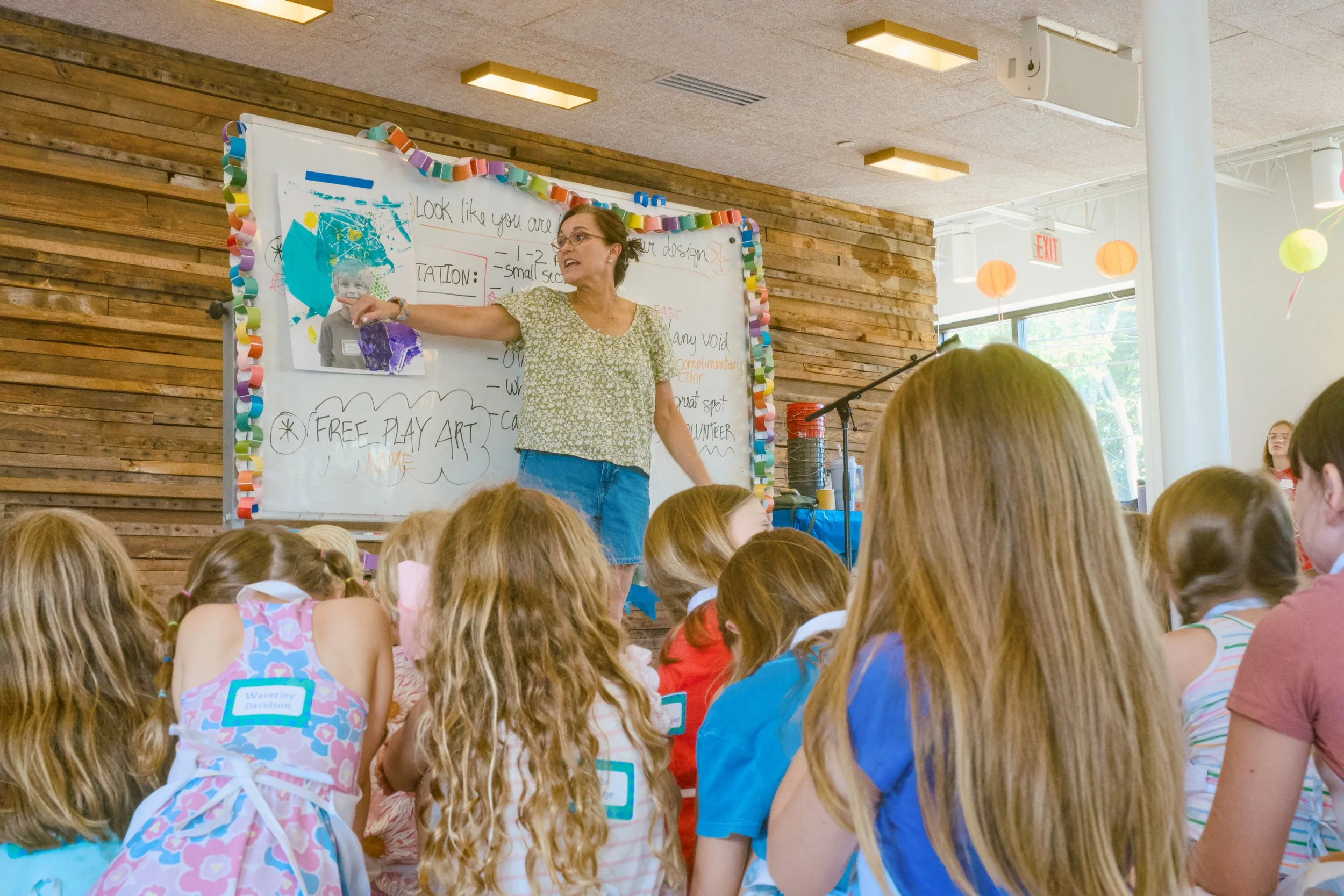 A woman teaching a class of children seated on the floor, with a whiteboard, colorful paper chain decorations, and a wooden wall in the background.