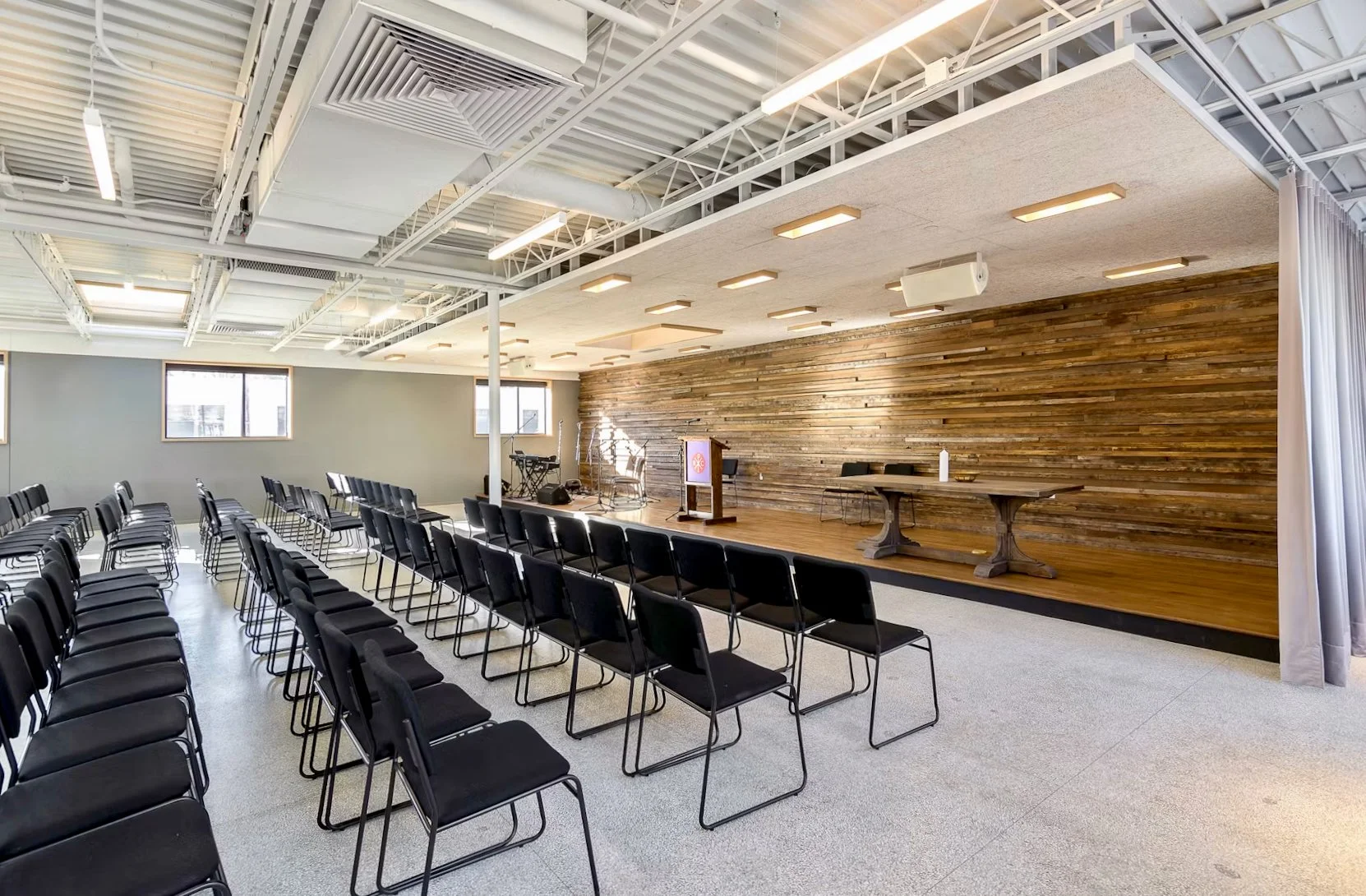 An empty conference room or event space with rows of black chairs facing a wooden stage with a podium and musical instruments, and a wood-paneled wall backdrop.