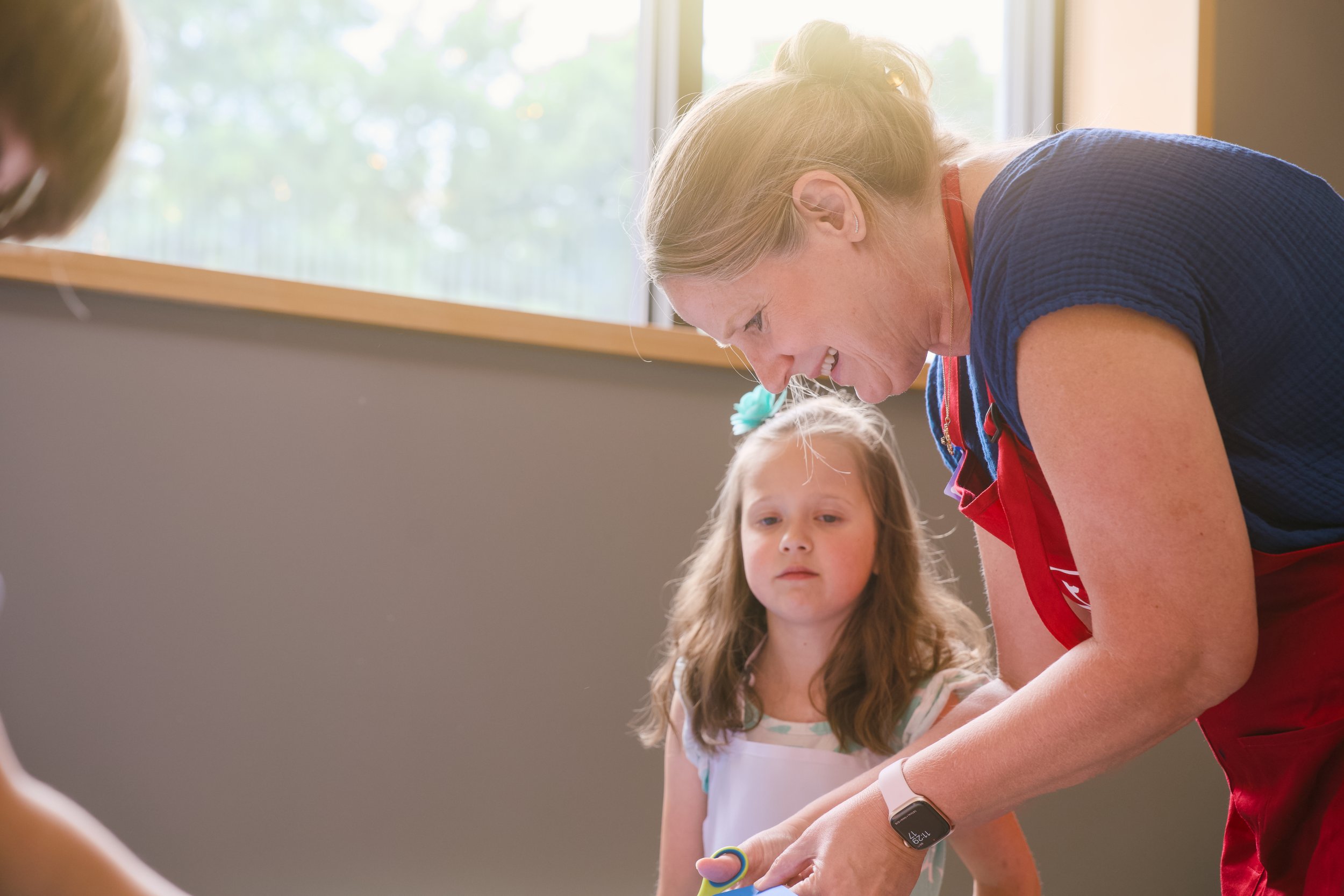 A woman with blonde hair tied in a bun laughing and leaning over a young girl with long brown hair, in a brightly lit indoor space with a large window in the background.