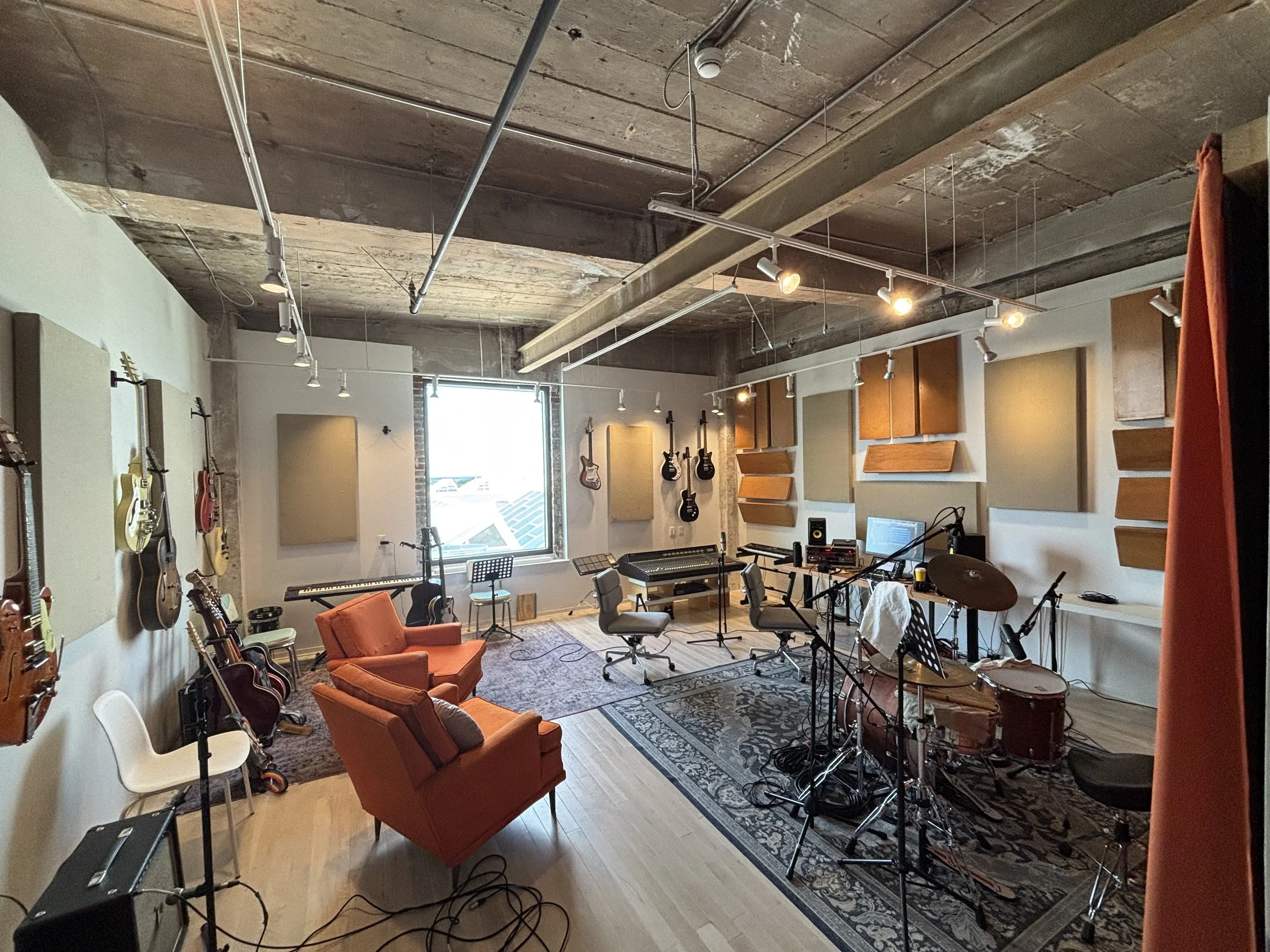Interior of a music recording studio with guitars hanging on the wall, musical instruments, soundproof panels, chairs, and recording equipment in a well-lit space with large windows and a decorative rug.