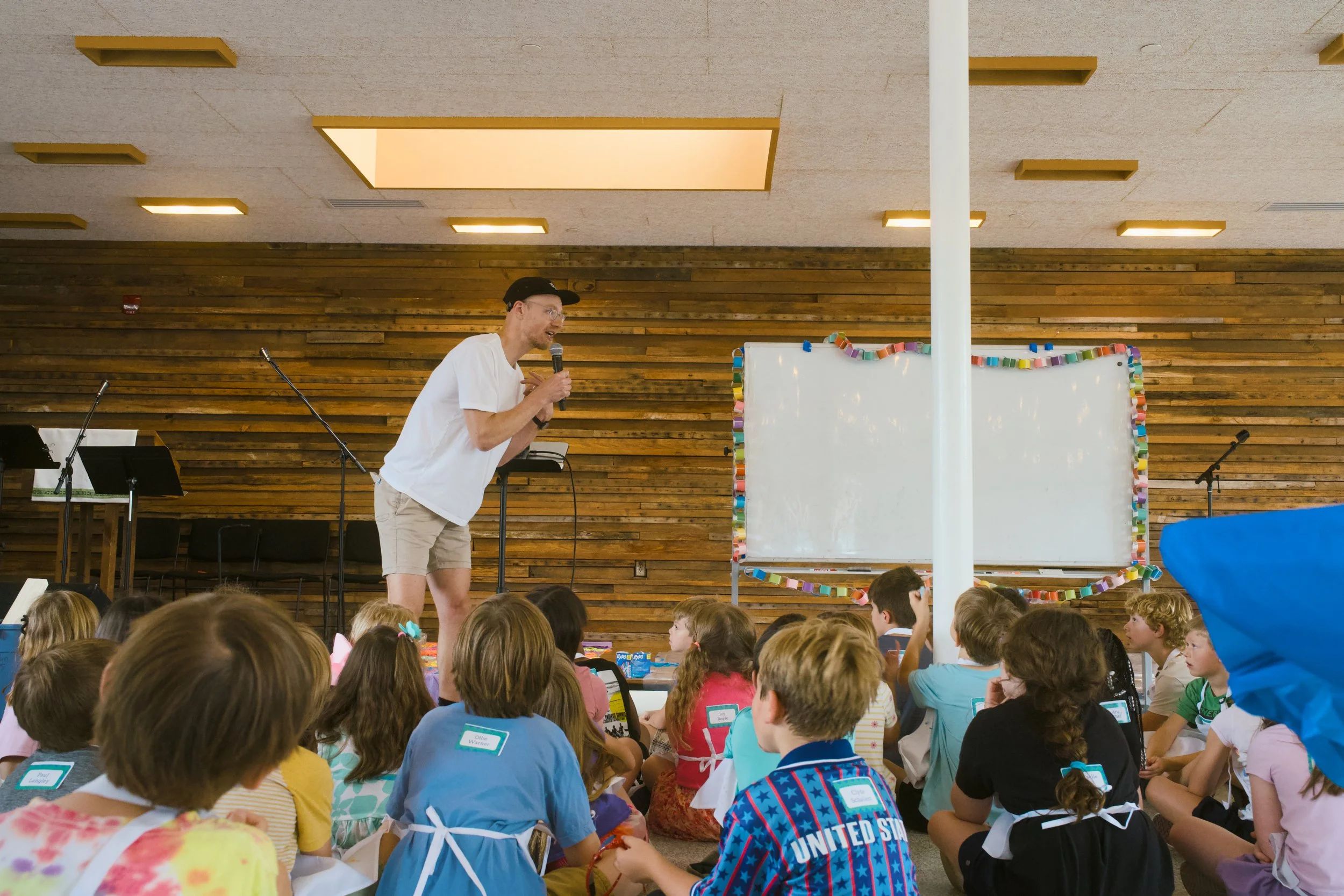 A man in a white shirt and beige shorts is speaking into a microphone in front of a large group of children seated on the floor, inside a room with wood-paneled walls and a decorated whiteboard.