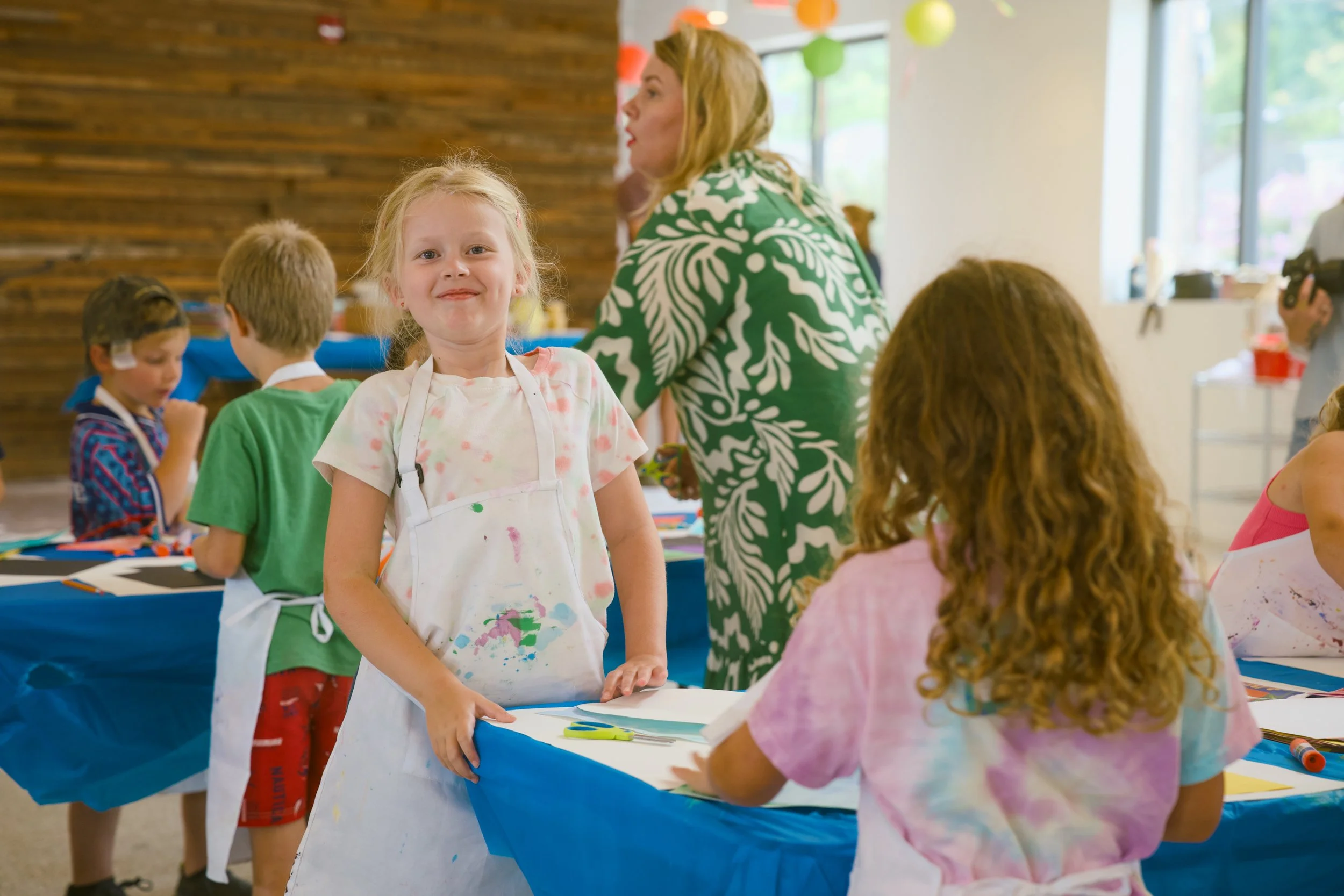 A young girl with blonde hair, wearing a painter's smock with paint stains, is standing in a brightly decorated room, smiling at the camera. Around her, children are engaged in art activities at tables covered with art supplies, and a woman is intera