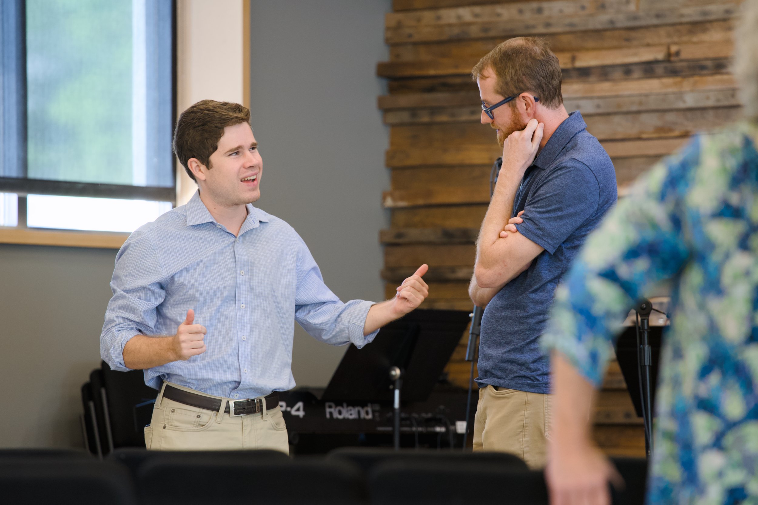 Two men are engaged in a conversation indoors. The man on the left is wearing a light blue shirt and beige pants, gesturing with his hands. The man on the right, wearing glasses and a dark blue shirt, is listening with his hand on his chin. There is a keyboard and wooden wall decor in the background.