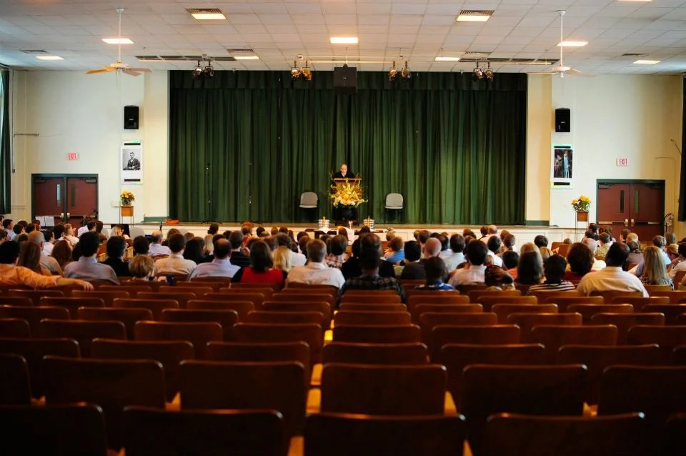 Audience seated in a theater watching a speaker at a podium on stage with green curtains.