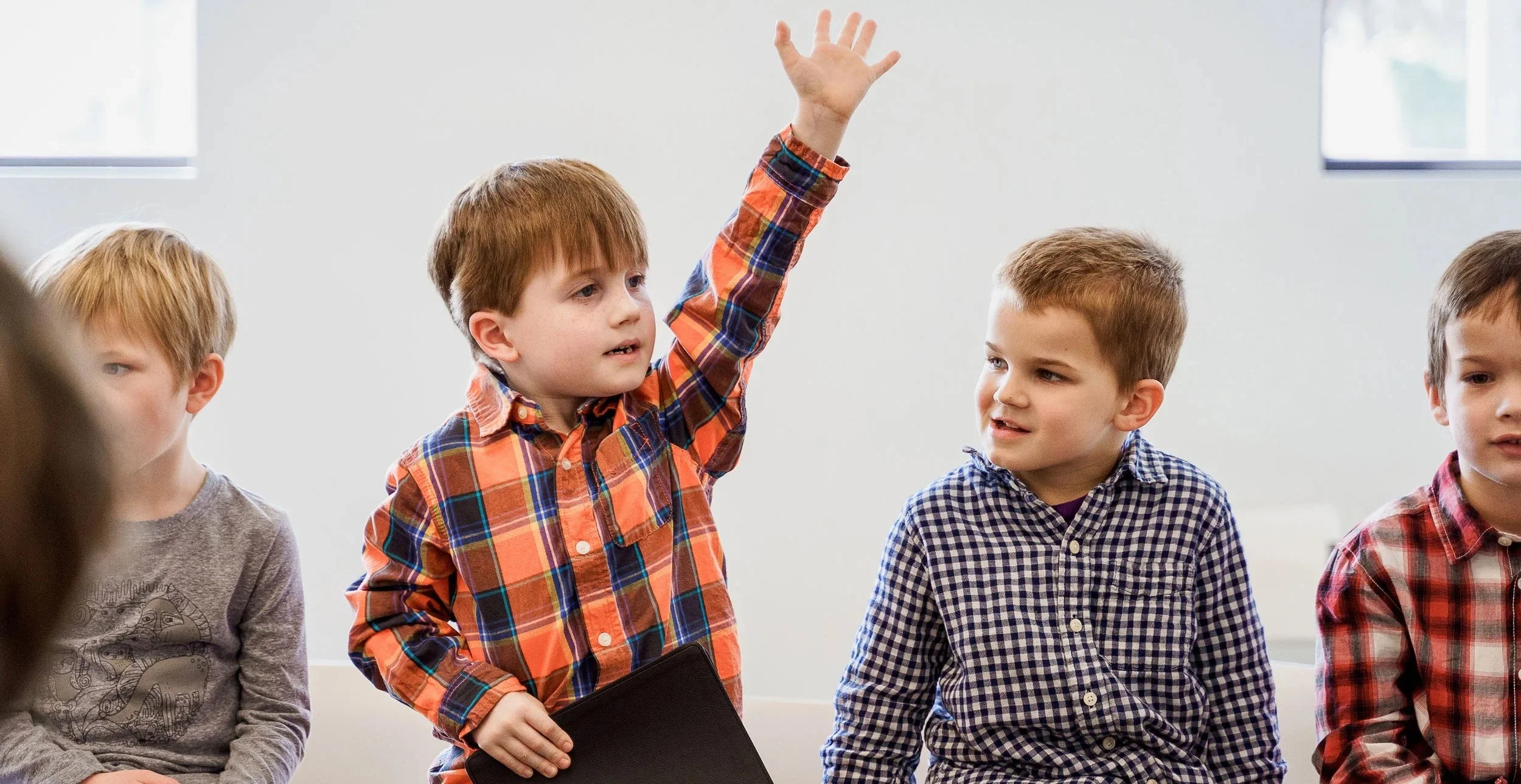 A group of young boys in a classroom, with one boy raising his hand to answer a question, while the others look on.