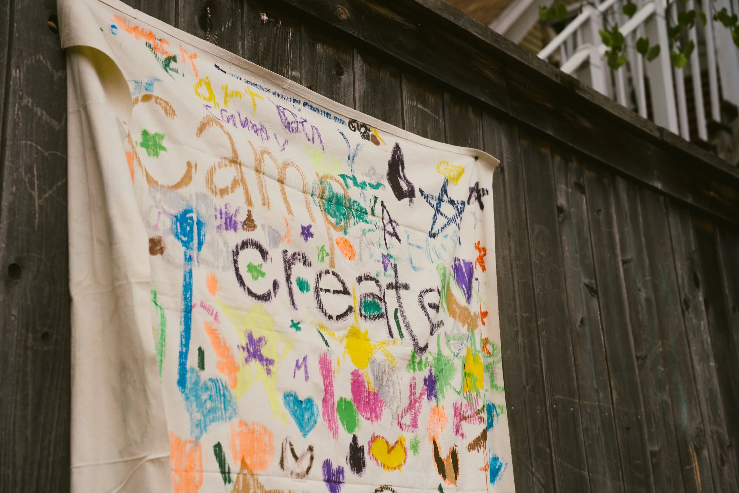 Colorful, child-like drawings and scribbles on a white cloth hanging on a dark wooden wall, with a second story white balcony in the background.