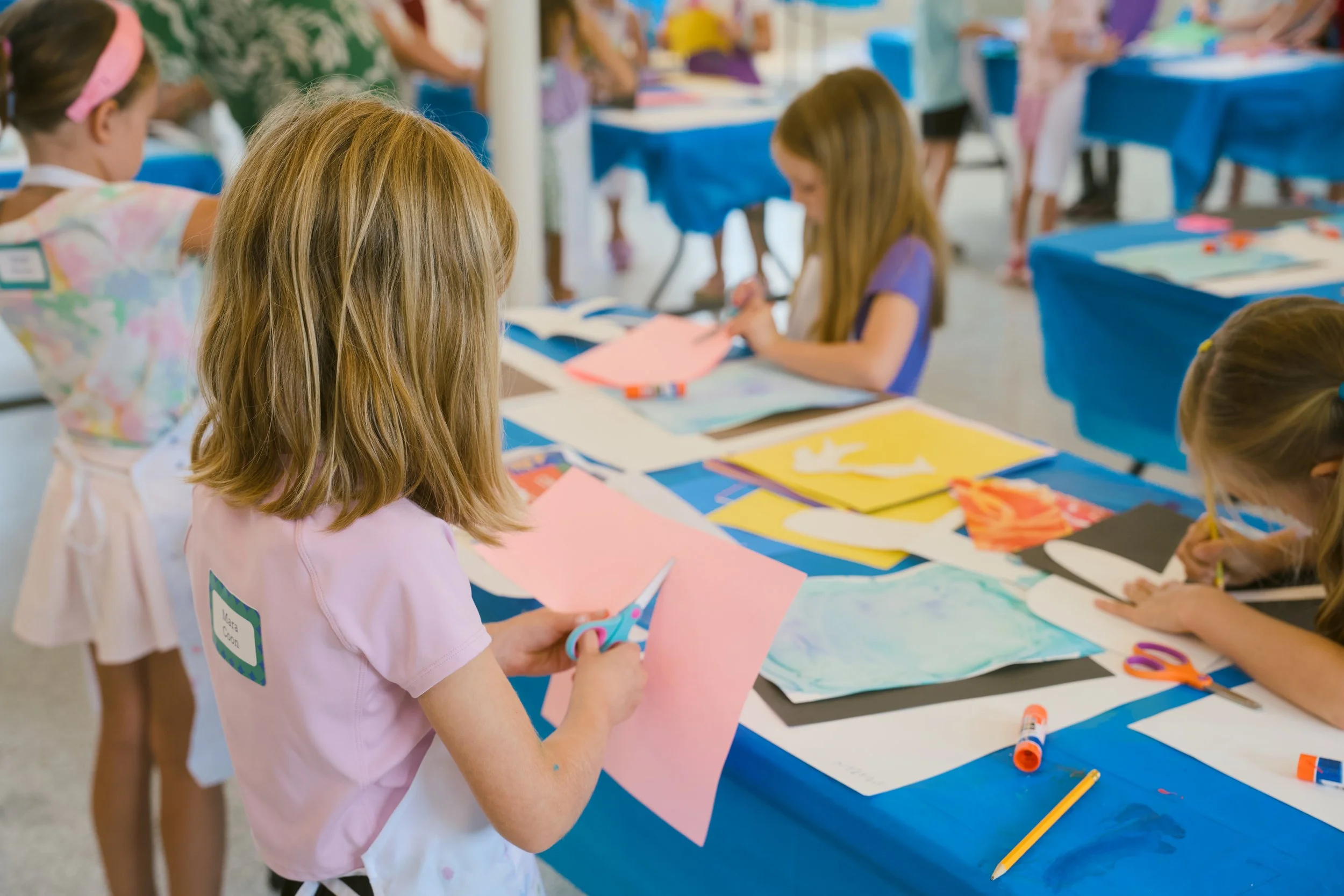 Children engaging in arts and crafts activity at a table covered with blue tablecloths, with colorful papers, scissors, and glue sticks, in an indoor setting.