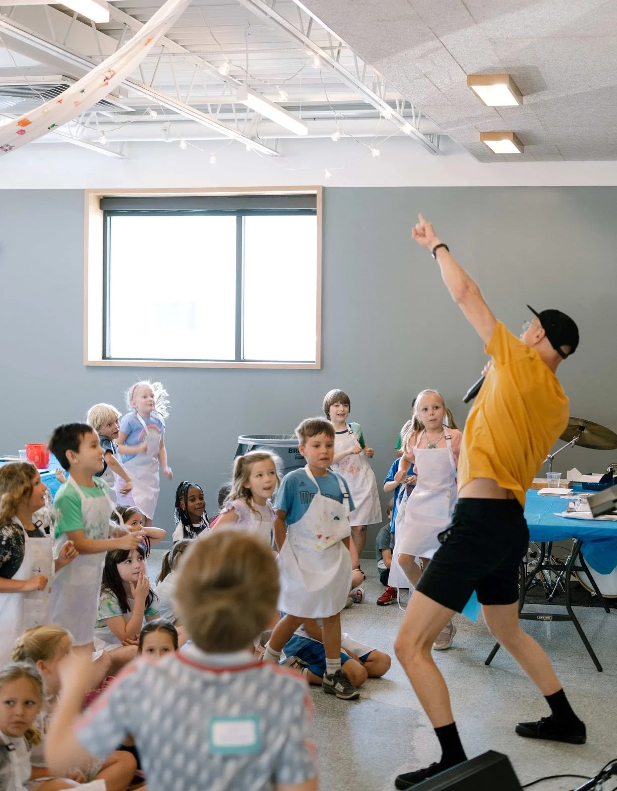 A group of children watching a performer in a yellow shirt and black shorts who is holding a microphone and pointing upward. The children wear aprons and appear engaged in a classroom or activity setting with a large window, gray walls, and ceiling l