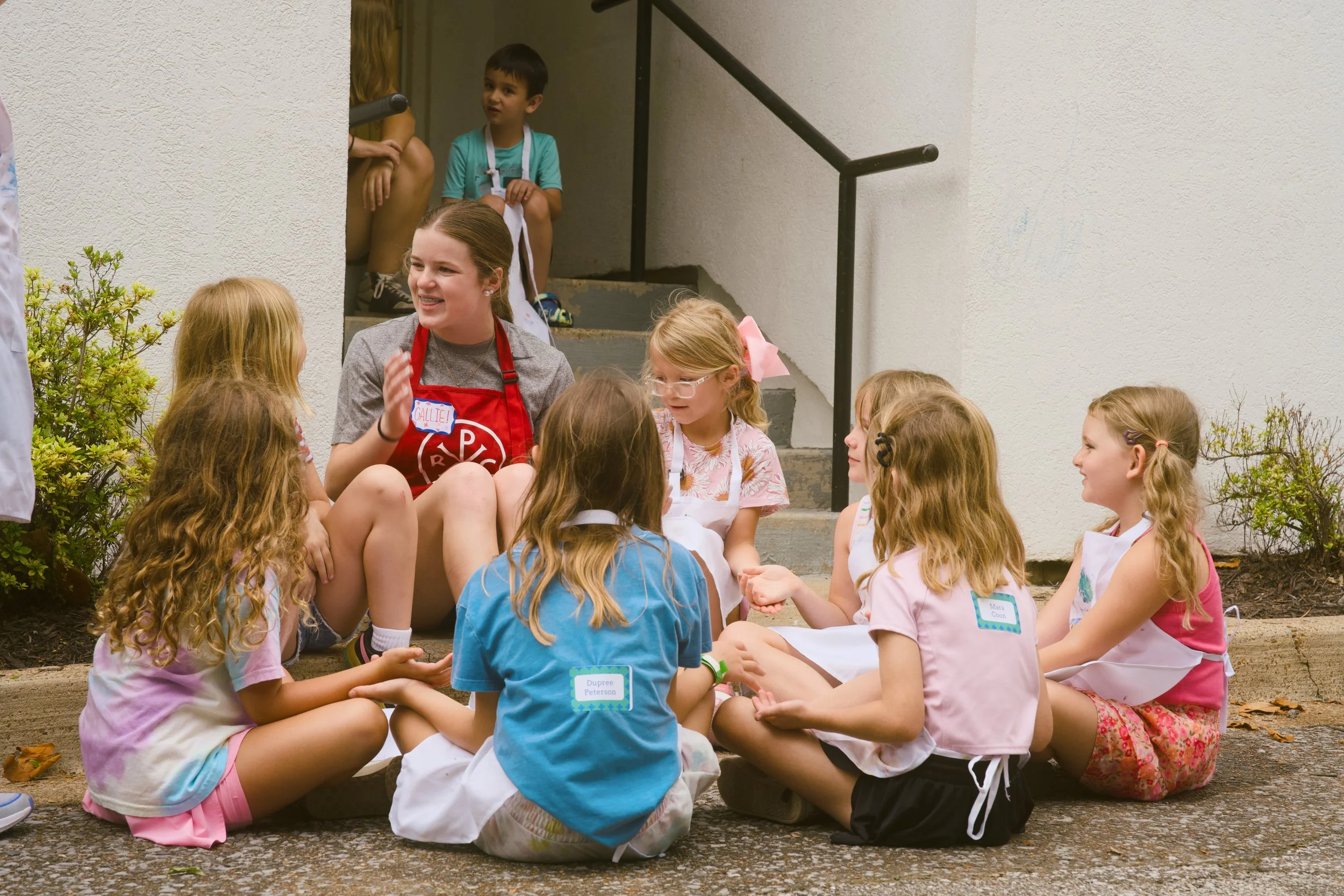 A group of children sitting in a circle outside, talking and laughing, with a young woman with a red apron sitting among them on the ground. A boy is sitting on stairs in the background with others, all engaging in conversation.