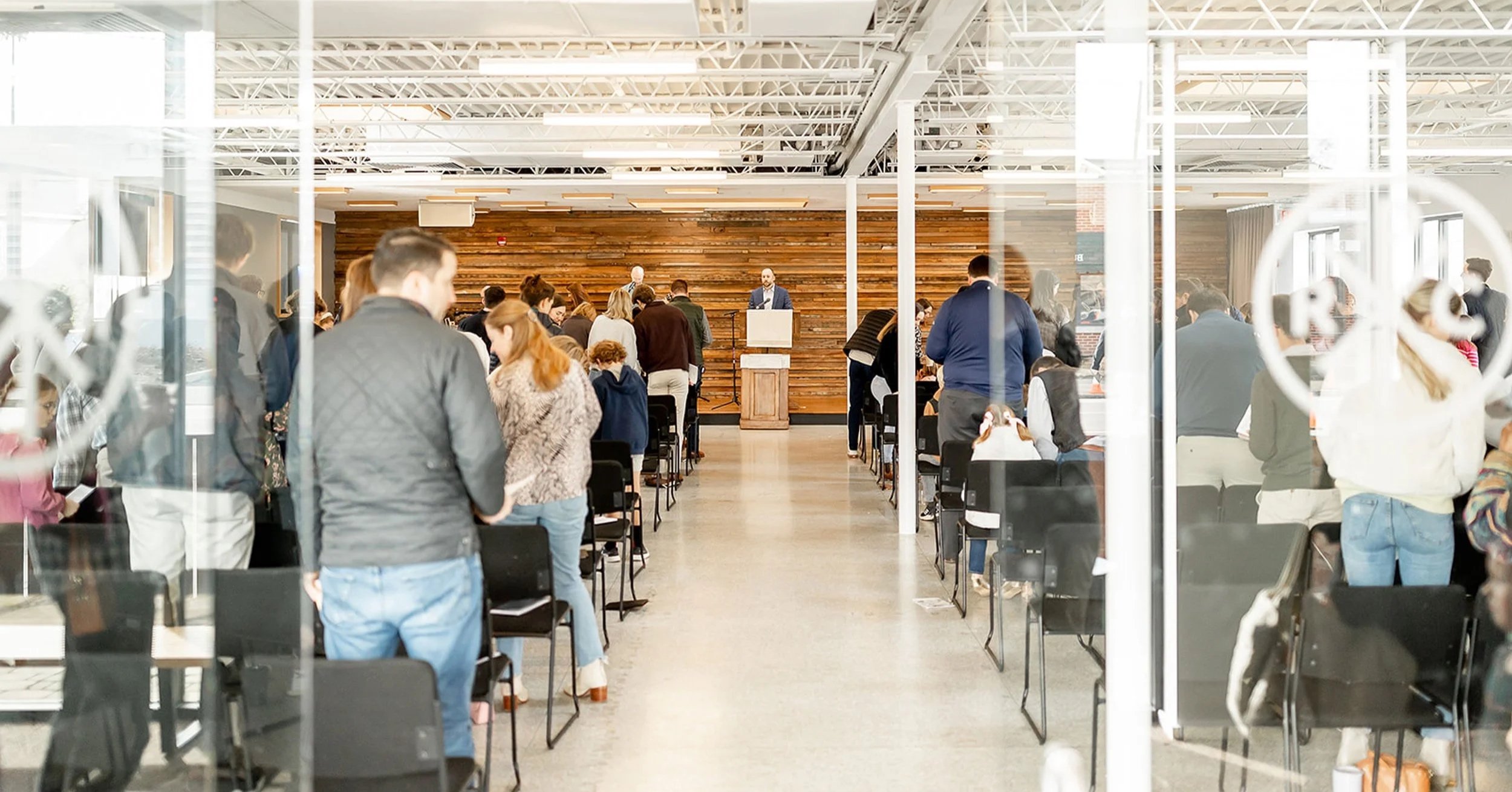 People standing and sitting in a modern church sanctuary with a wooden accent wall, while a speaker addresses the crowd from a podium at the front.