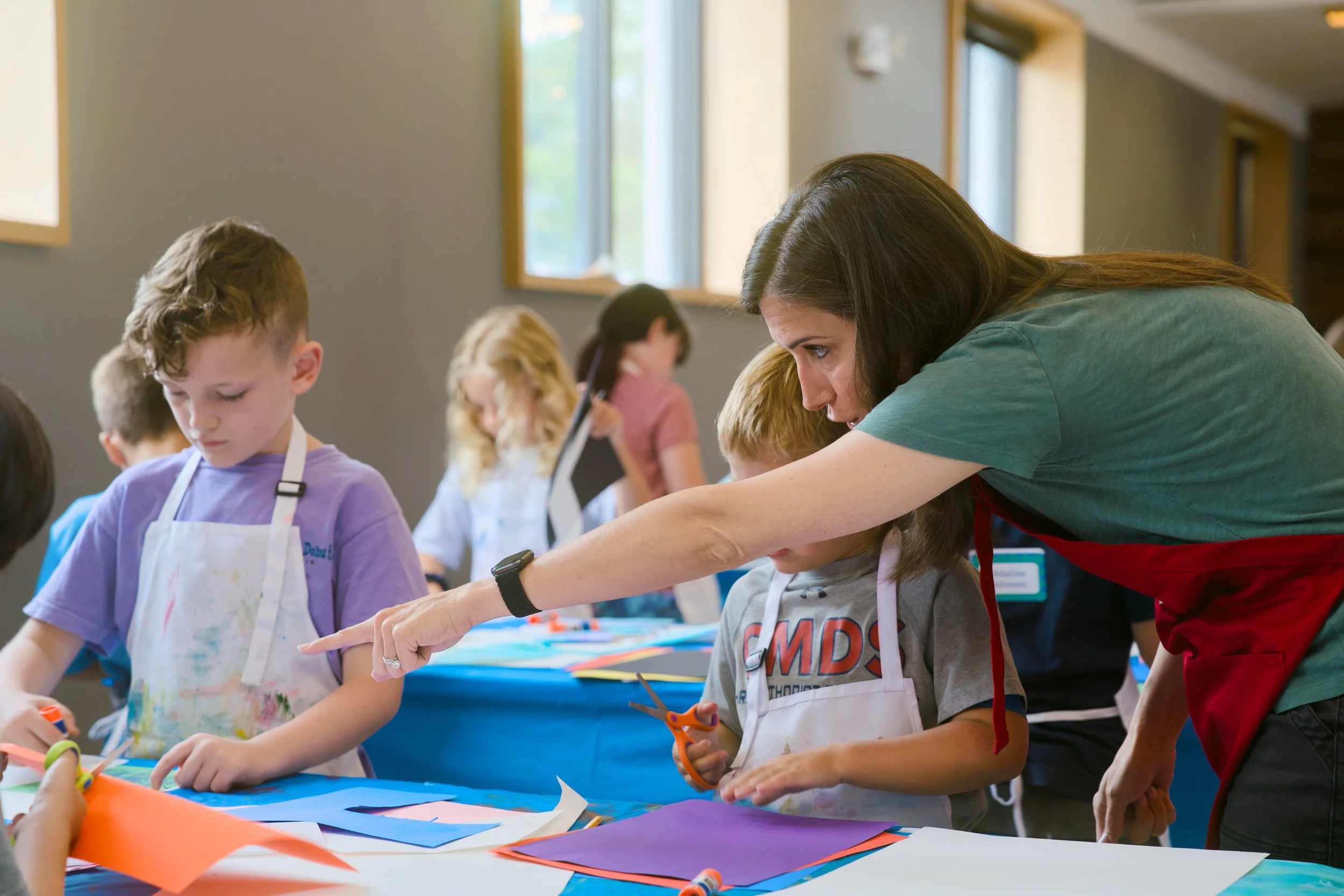 Teacher assisting children with arts and crafts project at a table with colored paper and scissors in a classroom.